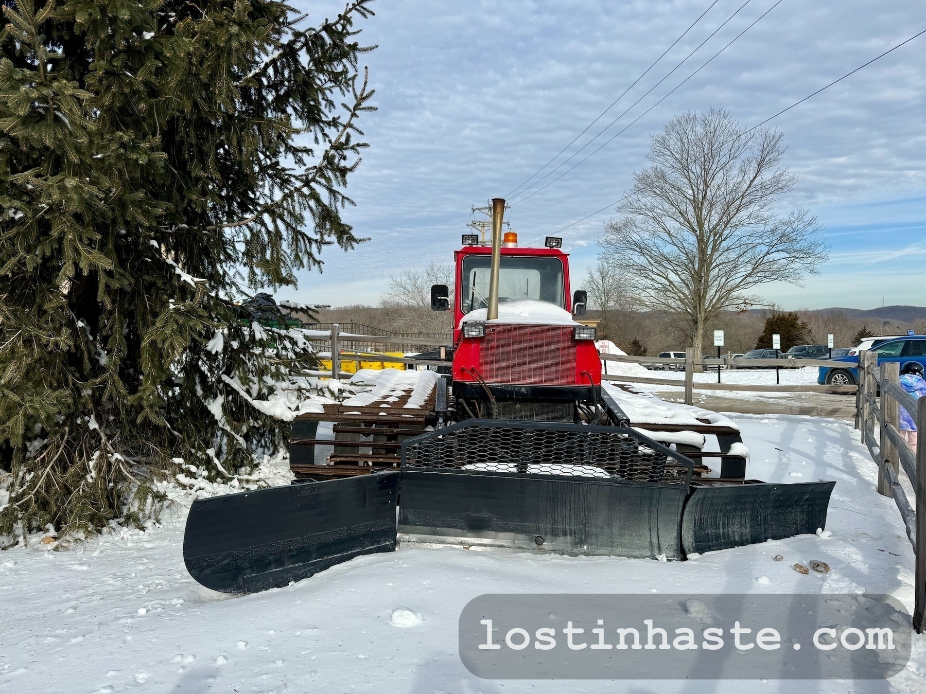 A red snow groomer with a front blade is parked on a snowy surface, near pine trees and a clear sky overhead.
