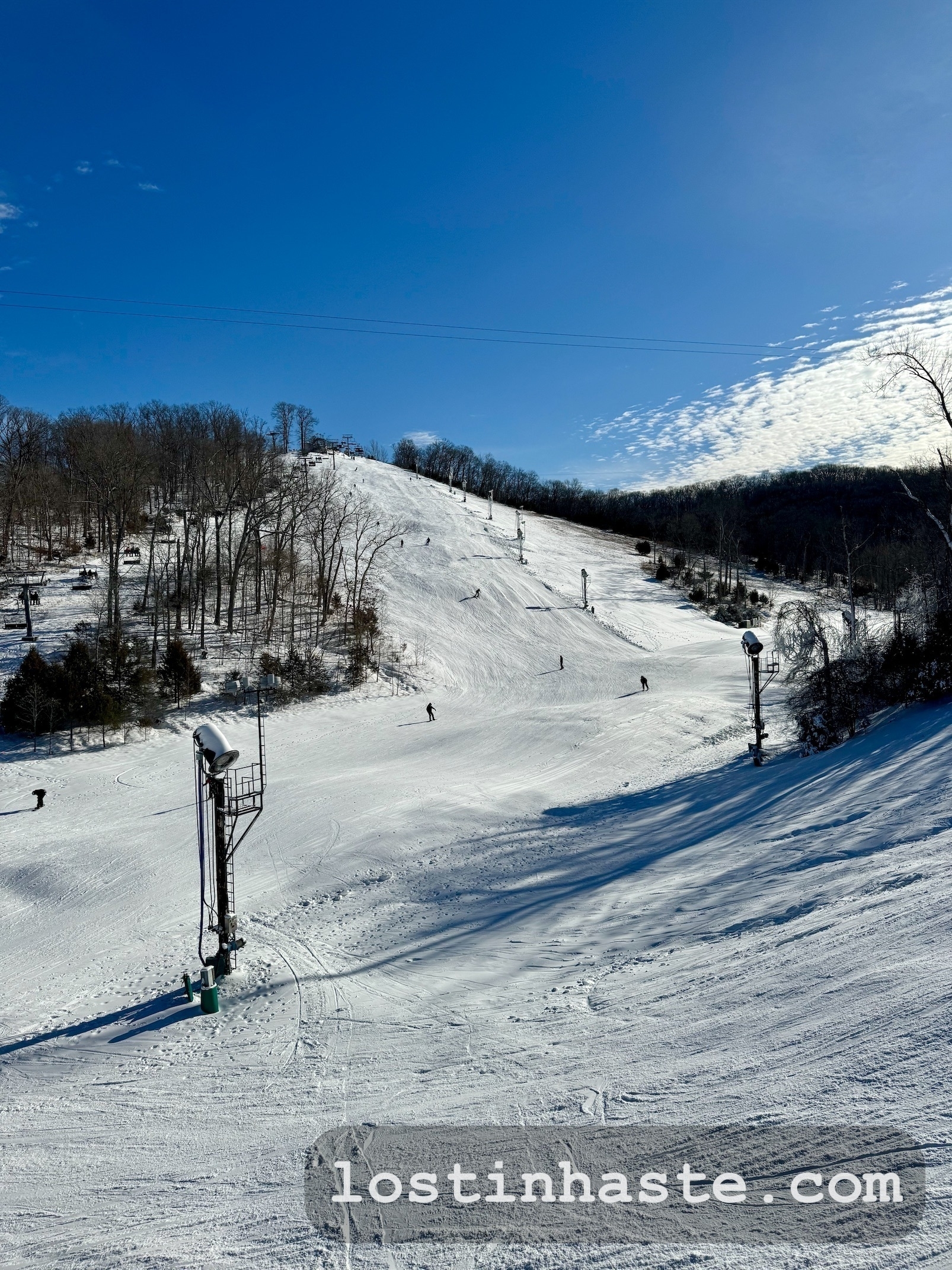 A snowy ski slope with skiers and a chairlift, surrounded by trees under a blue sky with wispy clouds.