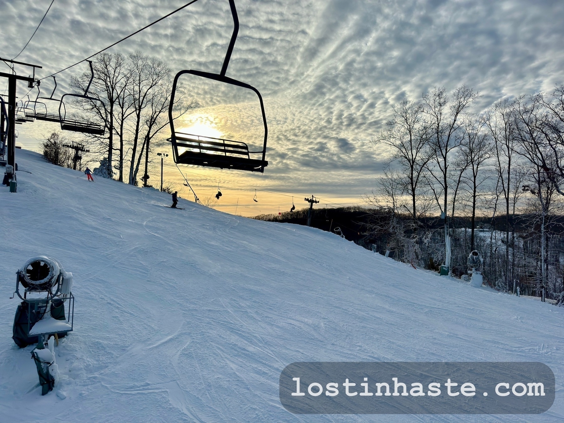 Ski lift chairs ascend above a snowy slope with skiers; textured clouds and a setting sun provide the backdrop.