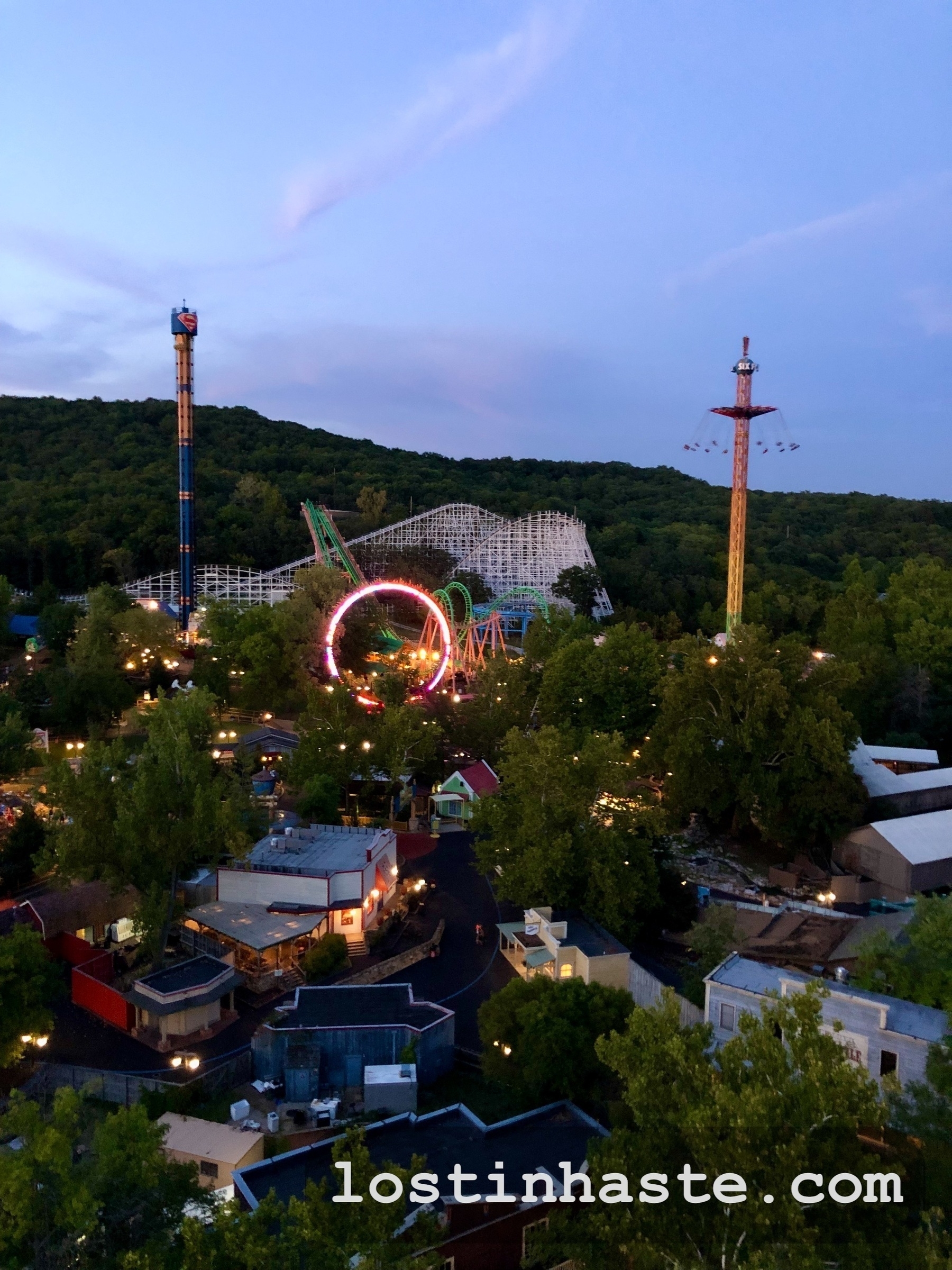 An amusement park at dusk featuring a lit Ferris wheel and roller coaster surrounded by trees.