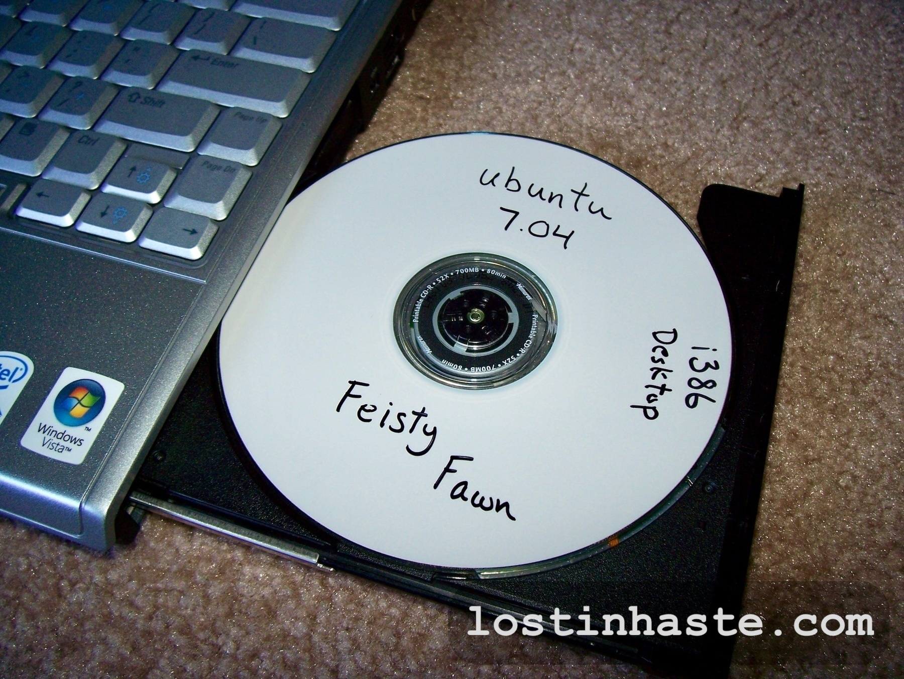 A CD labeled 'Ubuntu 7.04 Feisty Fawn Desktop i386' rests beside an open laptop on a carpeted floor. The website 'lostinhaste.com' is visible on the laptop.