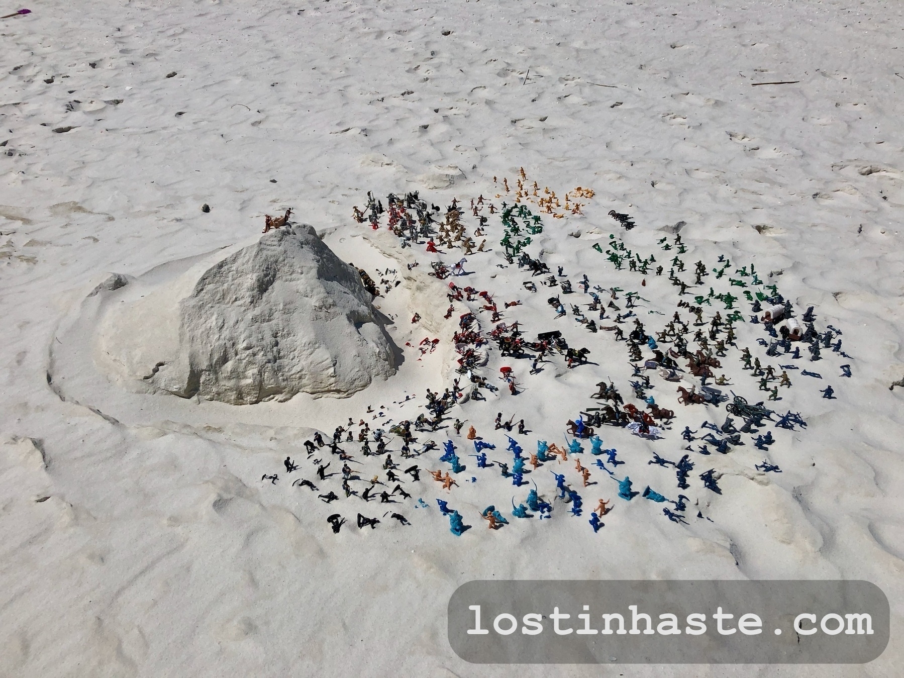 A large group of miniature figures is positioned around a grayish outcrop on a sand-covered ground, website address 'lostinhaste.com' displayed.