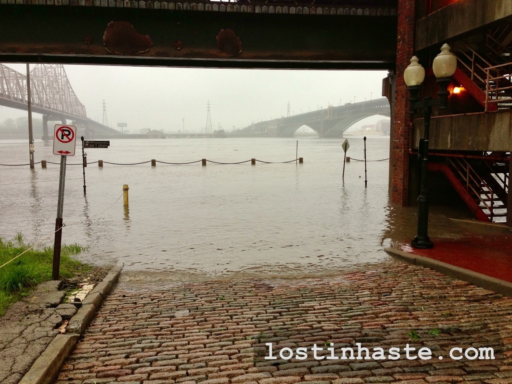 Floodwater covers a cobblestone street and sidewalk, with submerged traffic signs and street lamps, near bridges and buildings. Visible text: 'lostinhaste.com'
