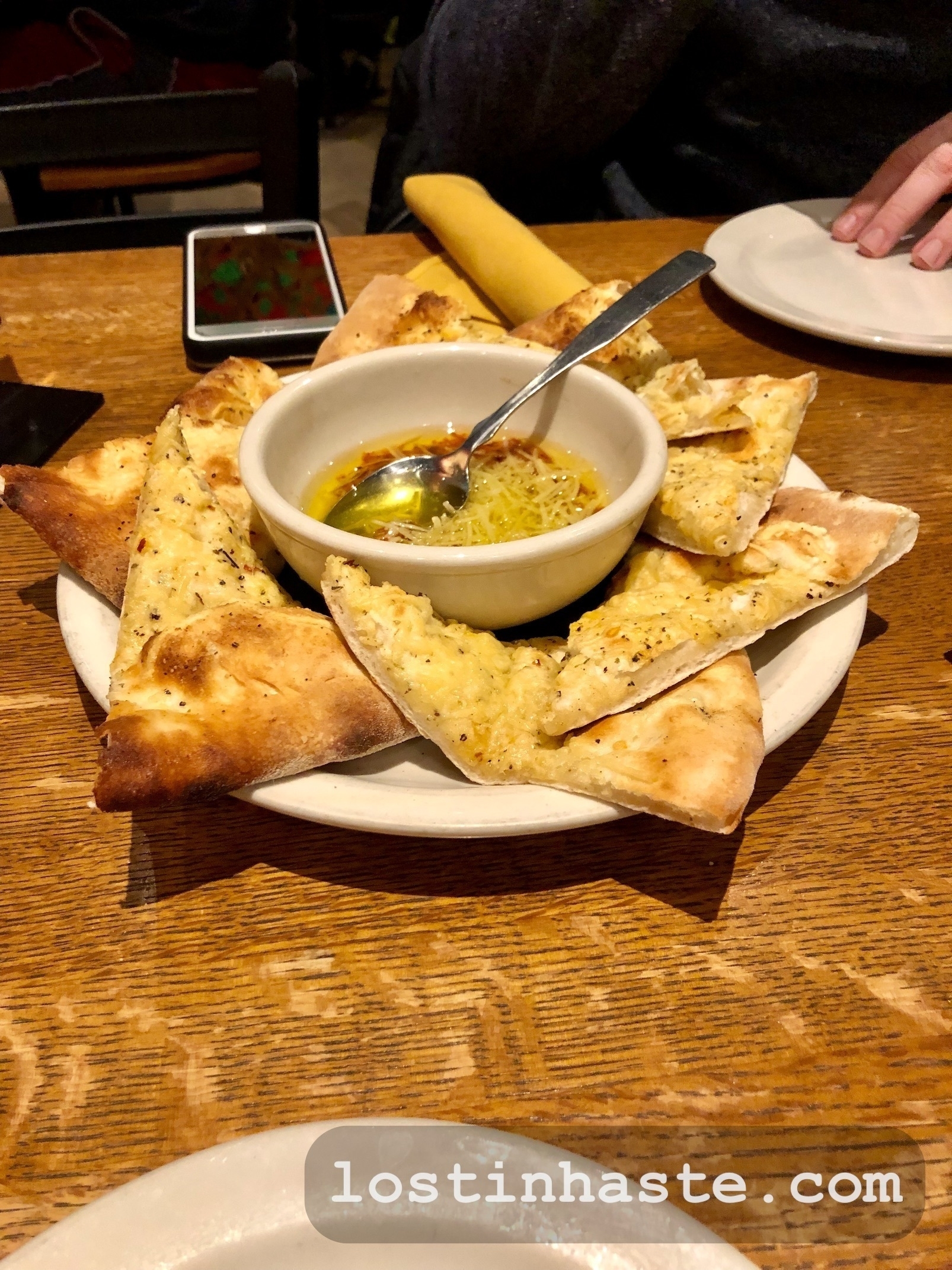 Triangular flatbread pieces surround a bowl of olive oil on a wooden table, suggesting a meal setting. Text at the bottom reads 'lostinhaiste.com'.