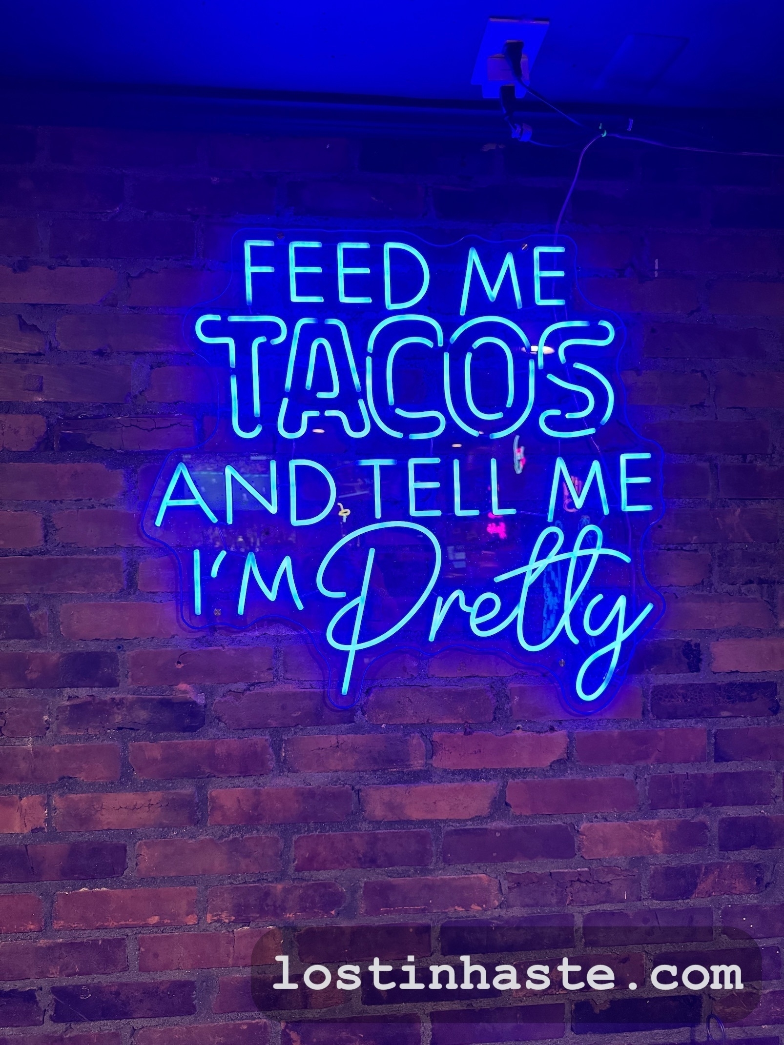 A neon sign on a brick wall displays the humorous message, FEED ME TACOS AND TELL ME I'M PRETTY, with a website URL at the bottom.