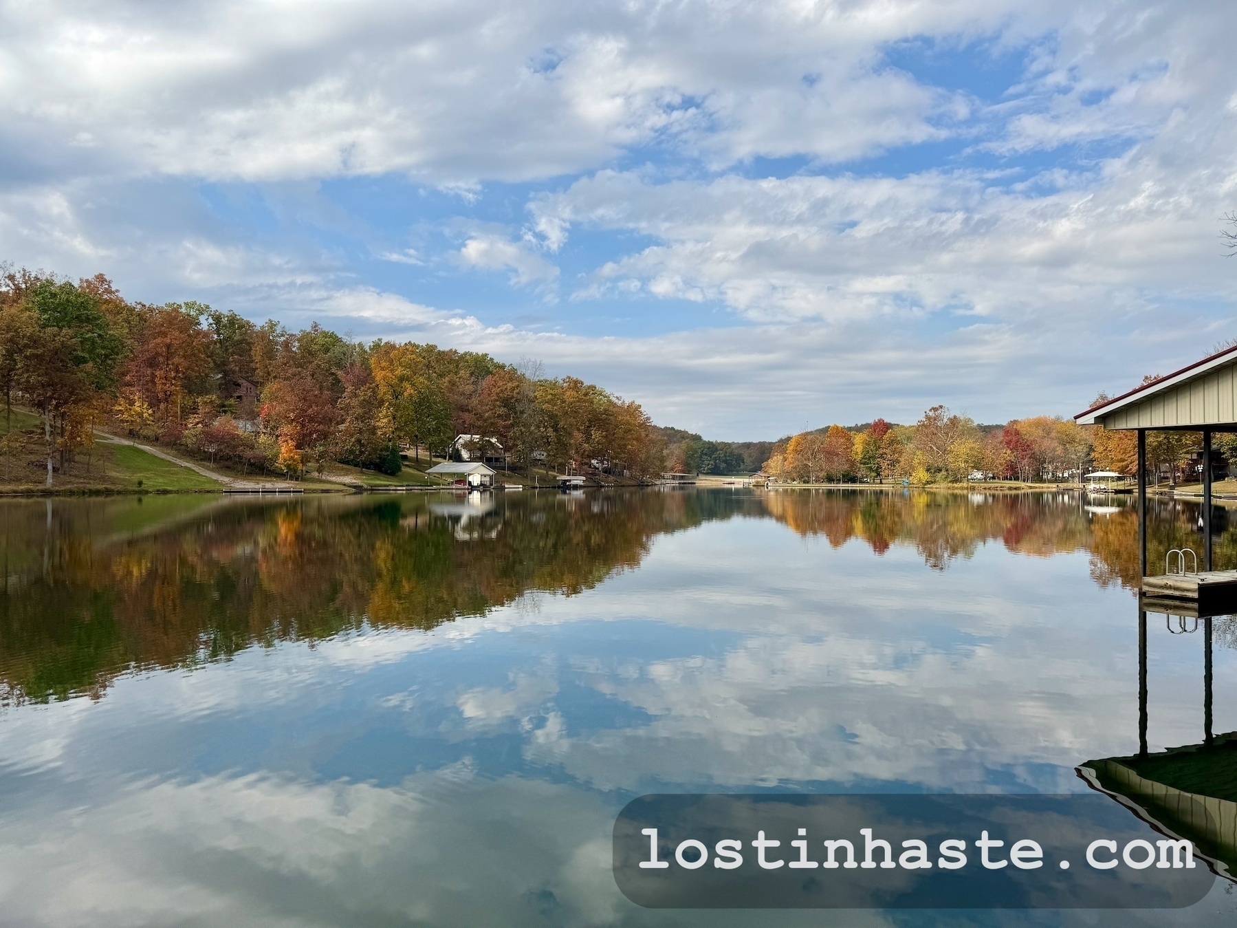 A serene lake reflects the autumn trees and sky, with houses lining the forested shoreline.