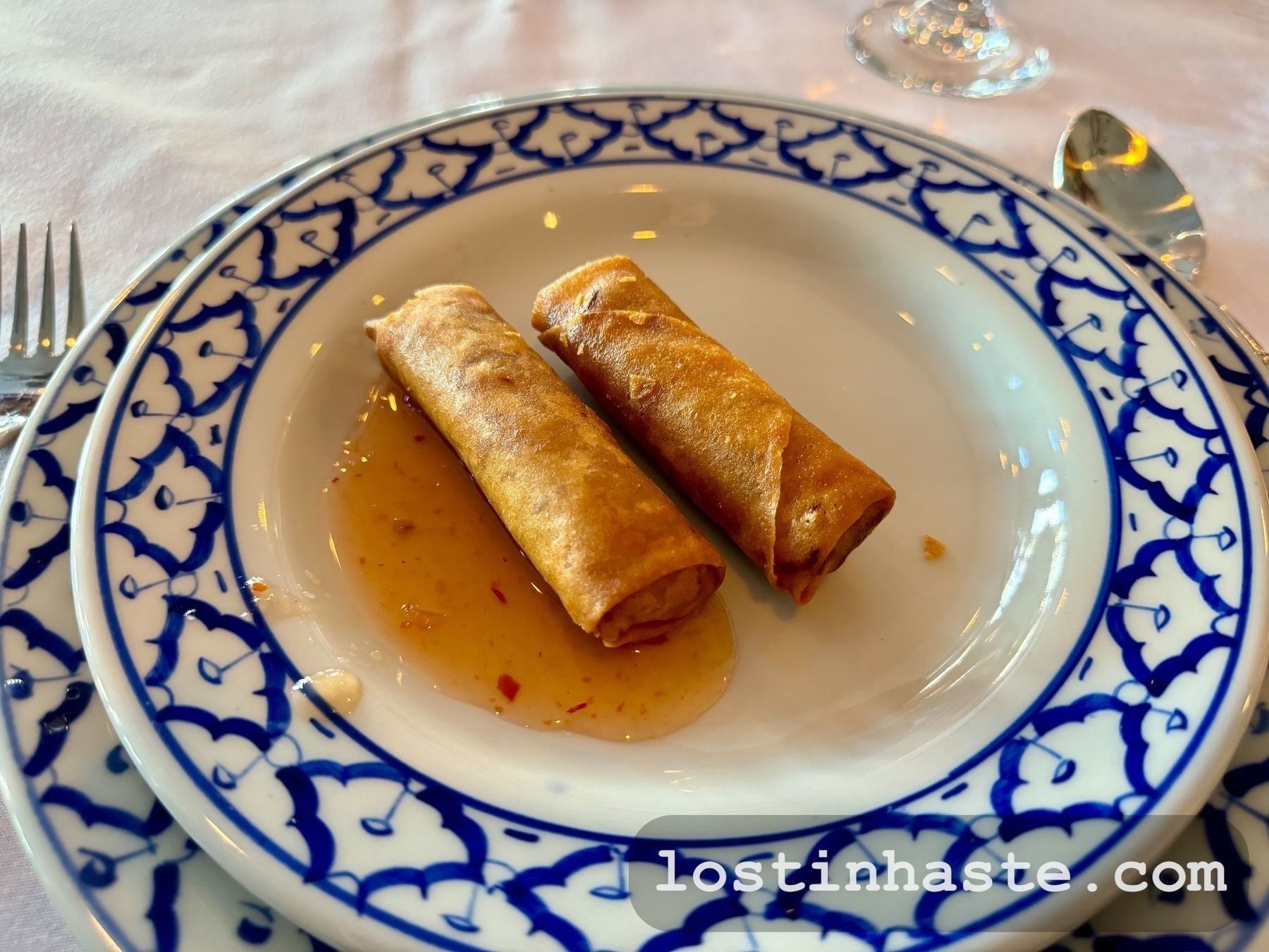 Two spring rolls sit on a detailed blue-and-white patterned plate with some dipping sauce, on a restaurant table.
