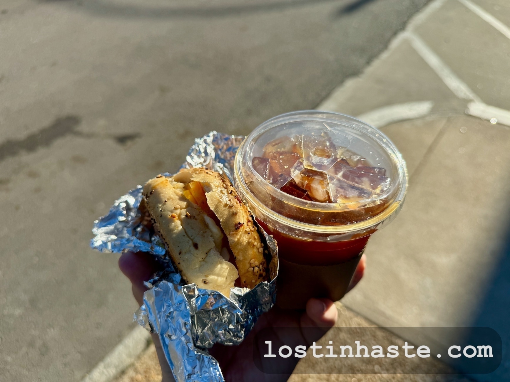 A hand holds a foil-wrapped egg bagel sandwich and an iced coffee against a blurred asphalt background. The text lostinhaste.com' is superimposed at the bottom.