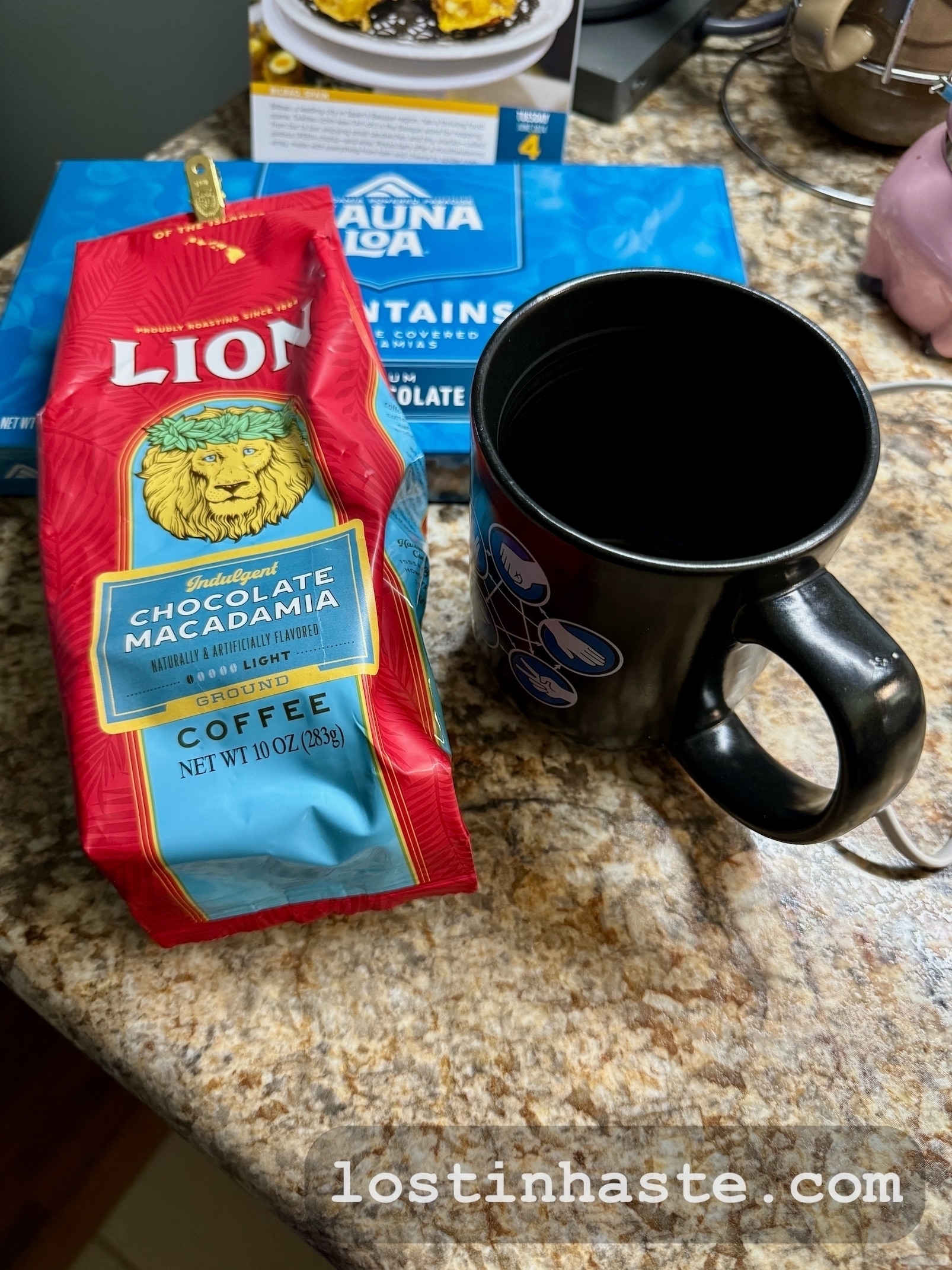 A red coffee bag labeled 'LION' rests on a countertop beside a Big Bang Theory (paper rocks lizard scissors Spock) themed mug and a blue box reading 'MAUNA LOA', all atop a kitchen counter. The URL 'lostinhaste.com' is overlaid at the bottom.