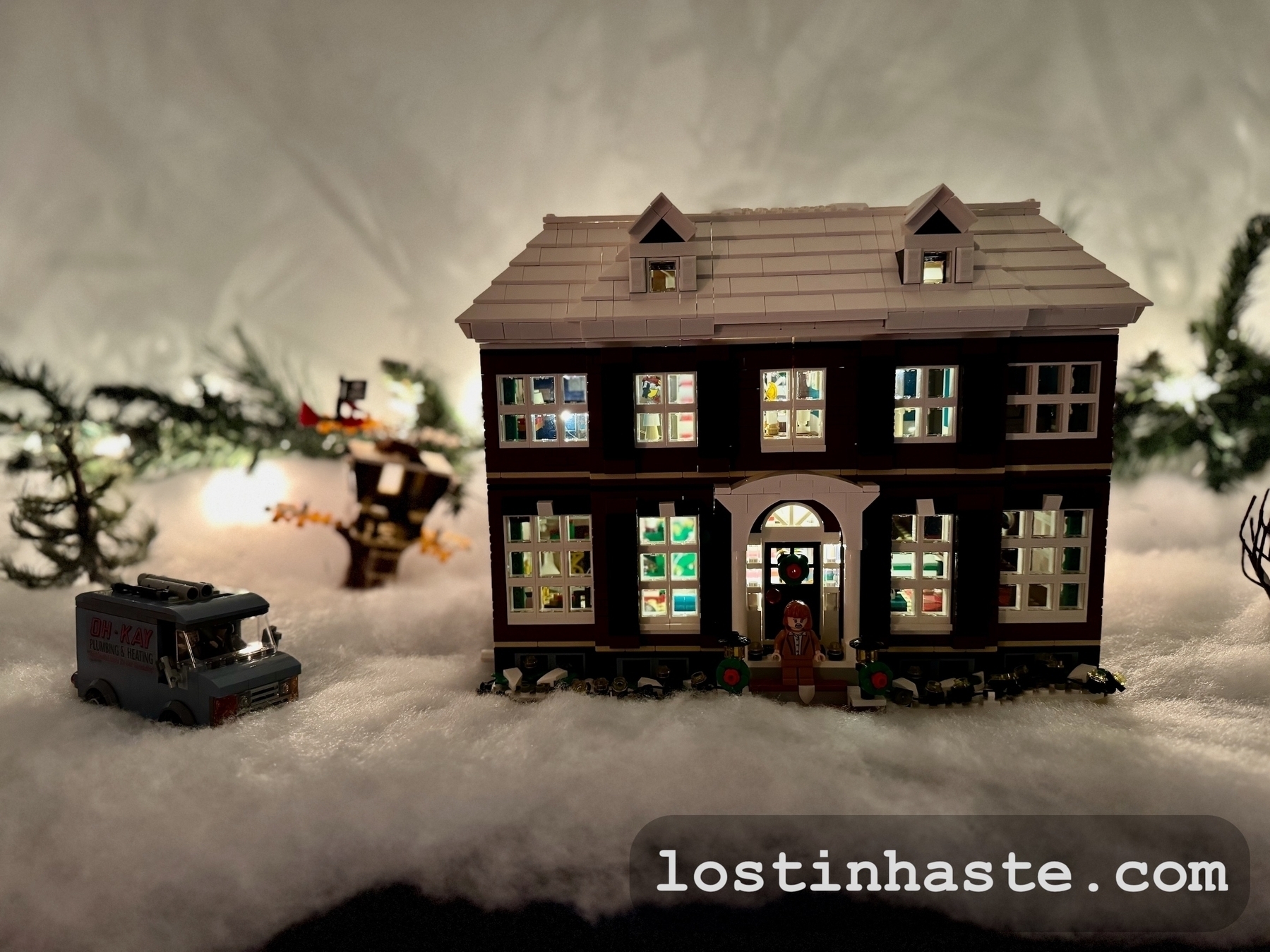 A miniature house (the Home Alone house, in Lego form), filled with lights, and van on a snowy landscape, surrounded by trees, with the website address 'lostinhaste.com' displayed as a watermark.