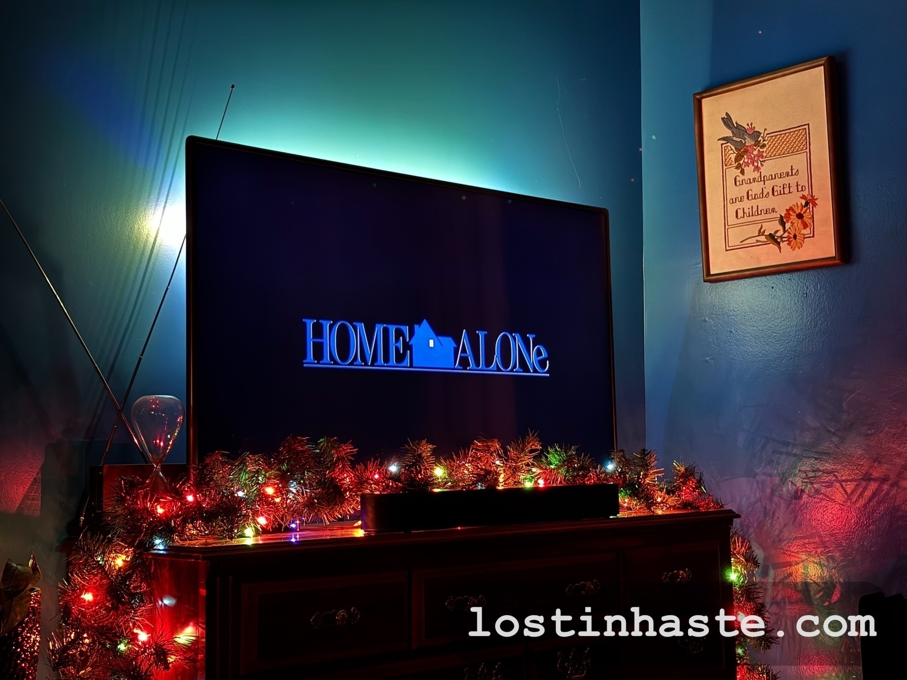 Auto-generated description: A TV displaying Home Alone is surrounded by colorful holiday lights in a cozy room with a framed picture on the wall.