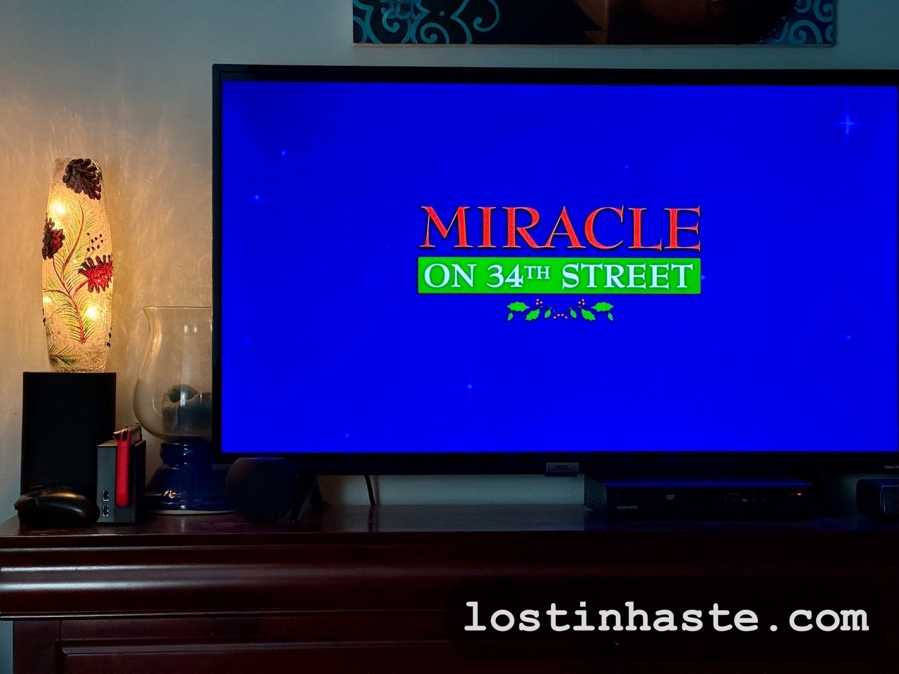 Auto-generated description: A TV screen displays the title Miracle on 34th Street with a decorative lamp on the left side and the website lostinhaste.com written at the bottom.