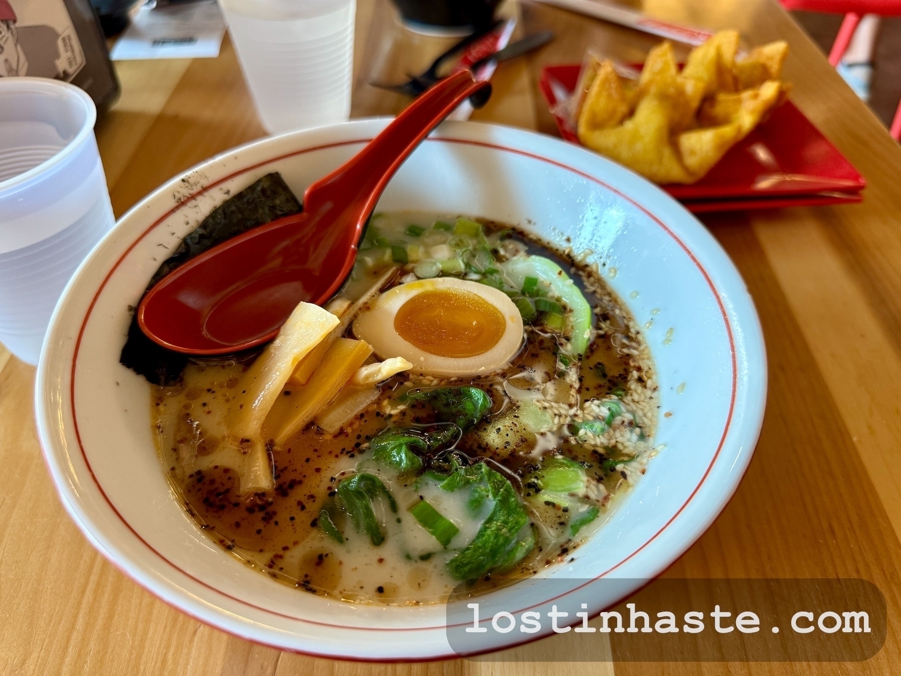 A bowl of ramen with toppings and a red spoon, set on a wooden table beside a side dish of gyoza and disposable cups; the watermark reads 'lostinhaste.com'.