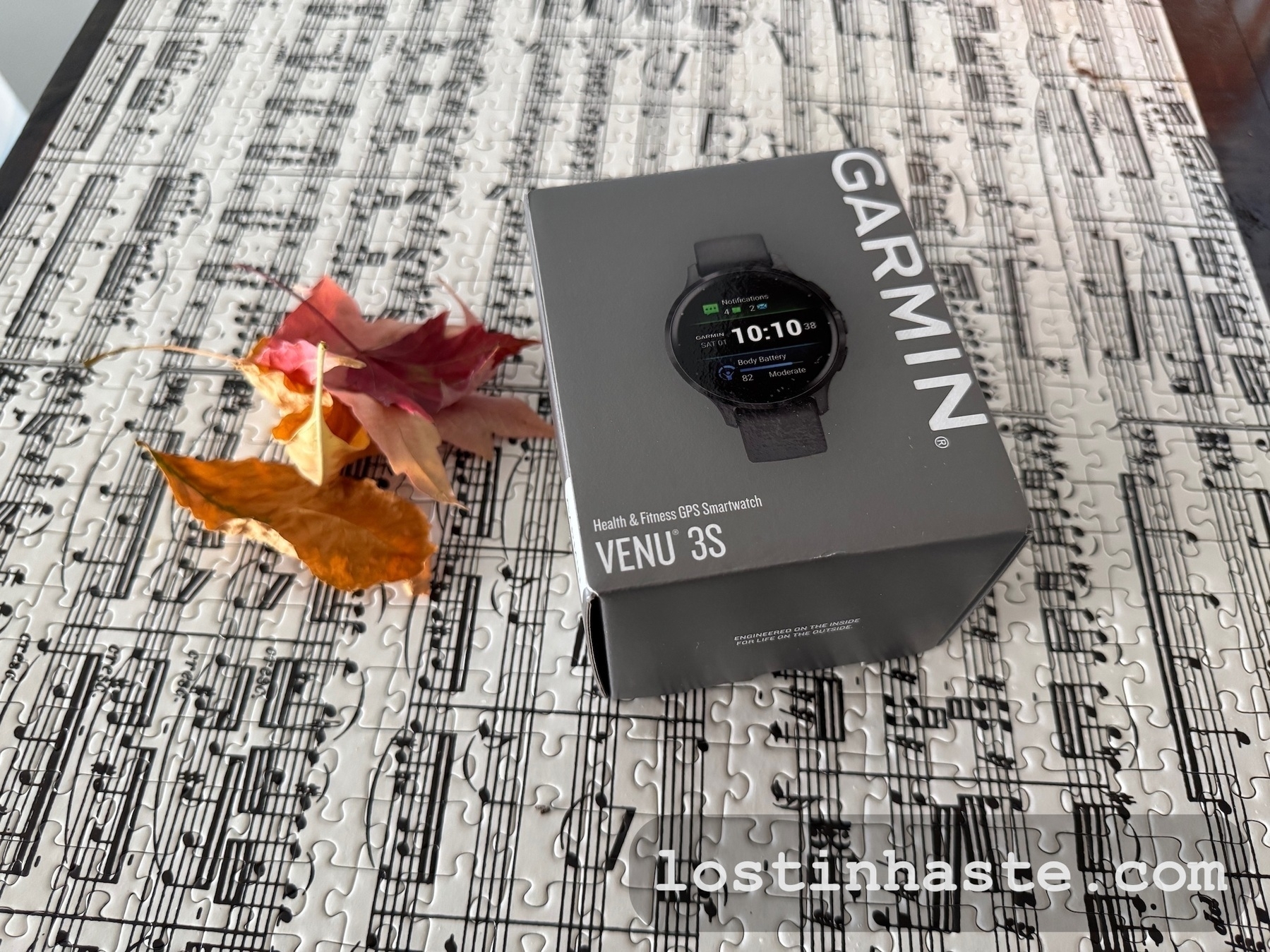 A Garmin Venu 3S smartwatch box is placed on a table with a puzzle and a few autumn leaves.