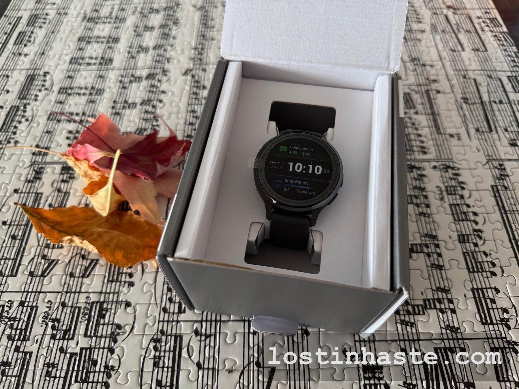 A smartwatch with a black strap is displayed in an open box on a table covered with a music-themed puzzle and accompanied by autumn leaves.