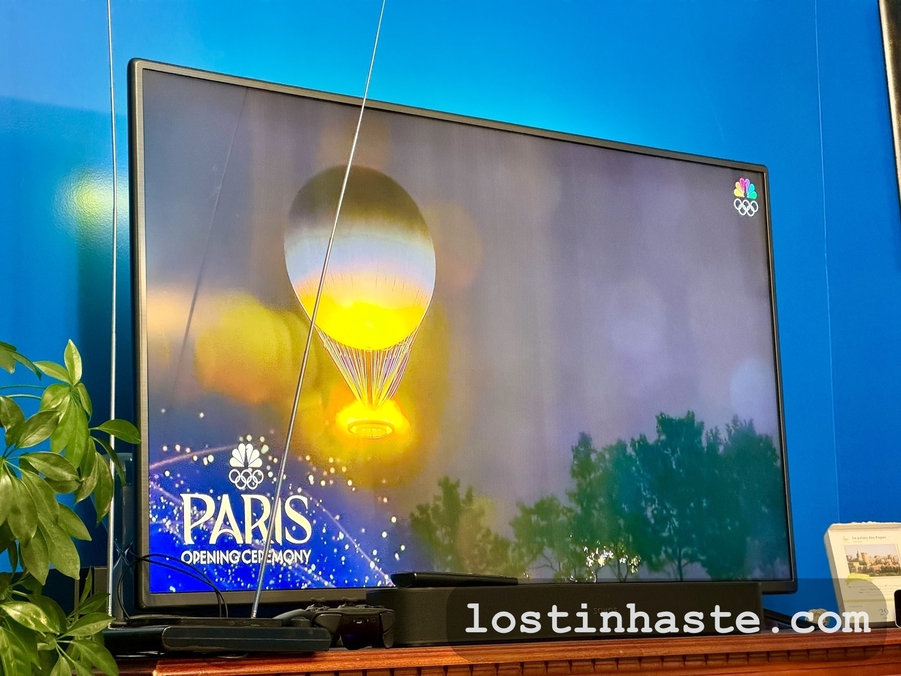 A television displays a hot air balloon over trees with 'PARIS Opening Ceremony' and the website 'lostinhaste' overlaid. Olympic rings and the NBC logo are in the corner.