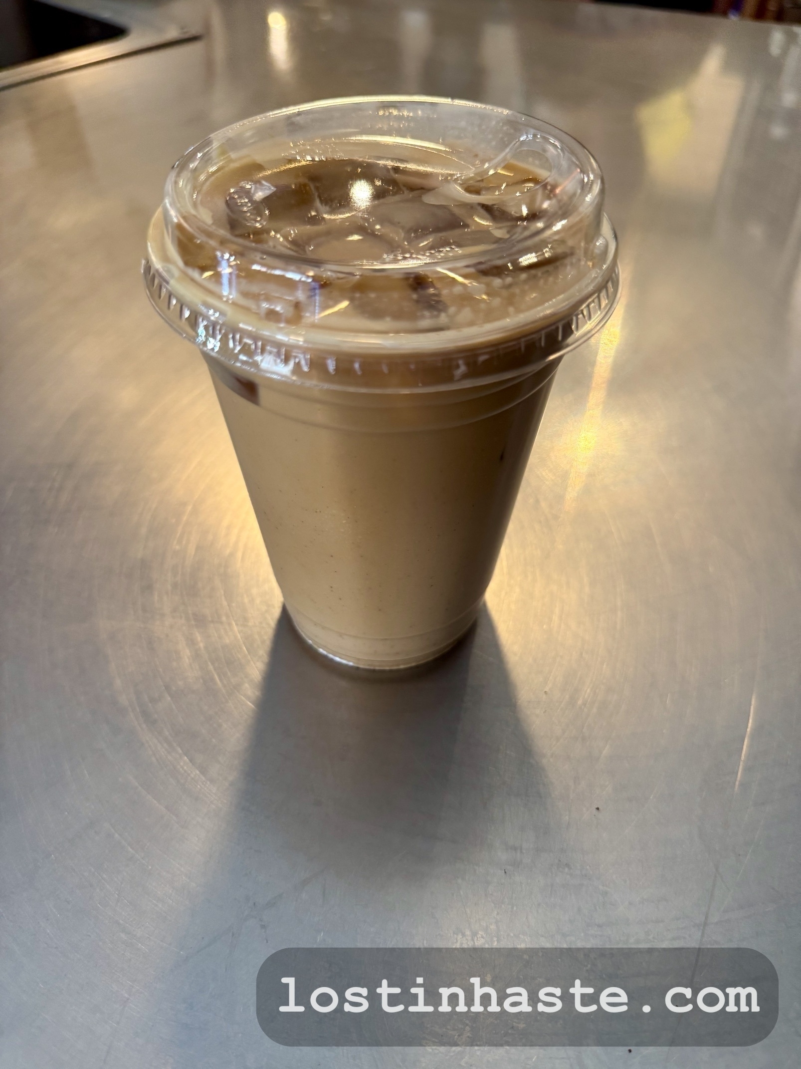 A plastic cup filled with iced coffee and a clear lid sits on a metallic surface, with the URL lostinhaste.com displayed on the image.