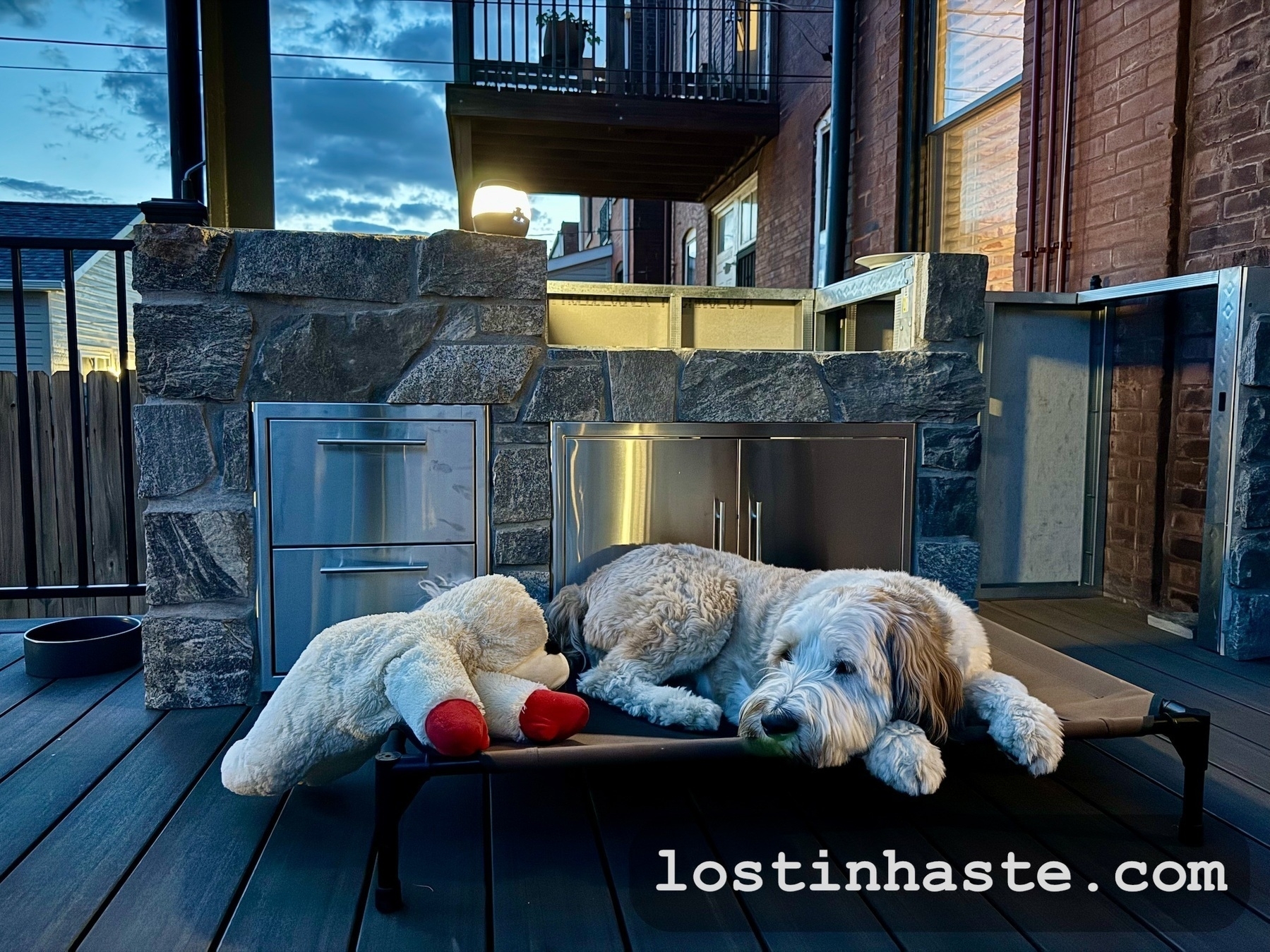 A dog reclines on an outdoor pet bed next to a plush toy, on a deck with stone-built structures and fencing, at dusk.