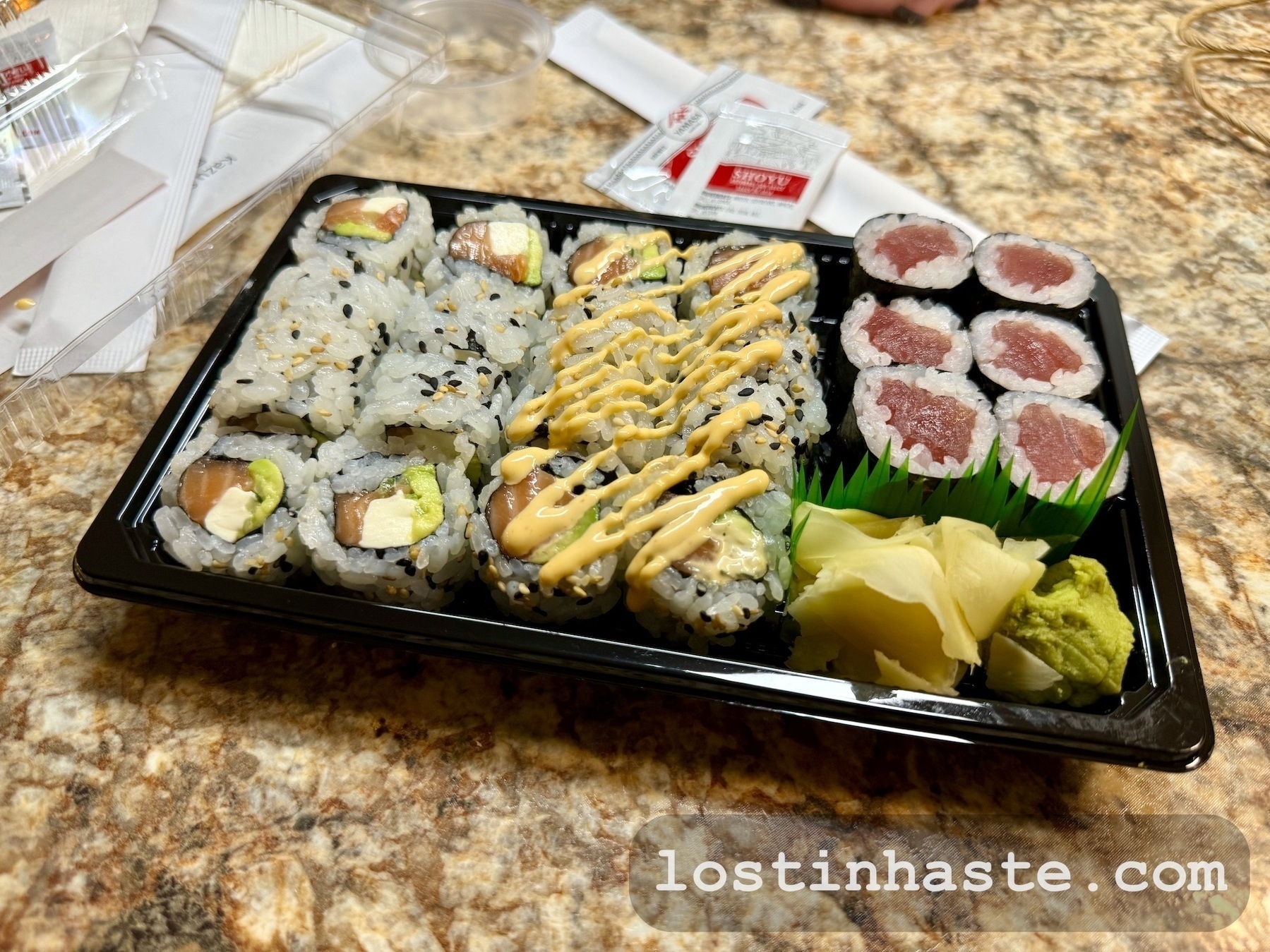 Assorted sushi in a plastic tray featuring rolls with avocado, fish, and drizzled sauce, alongside ginger and wasabi, on a kitchen counter with packets and a website URL: lostinhaste.com.