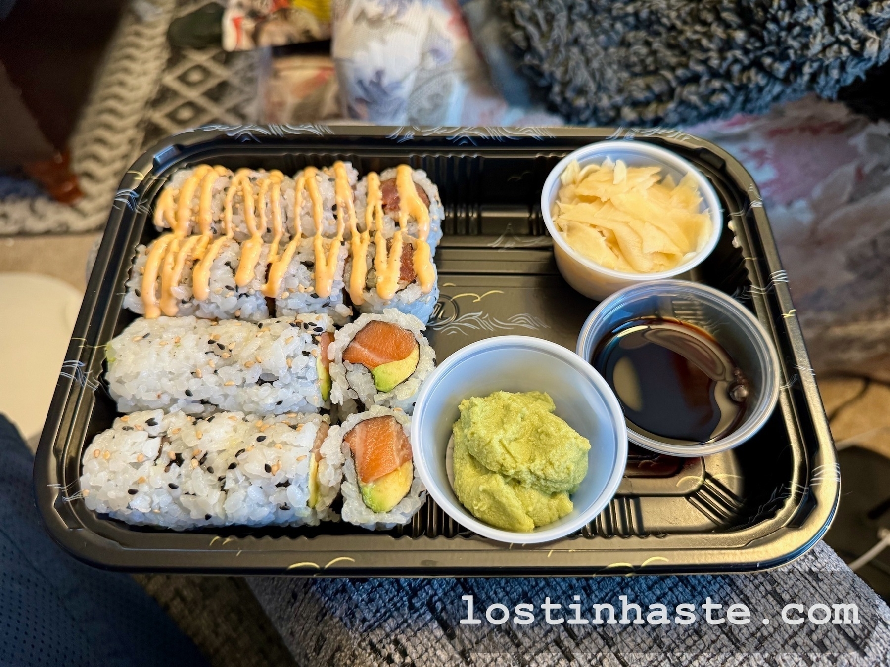 Sushi rolls in a tray with condiments; wasabi, pickled ginger, and soy sauce on the side, on a patterned fabric background.