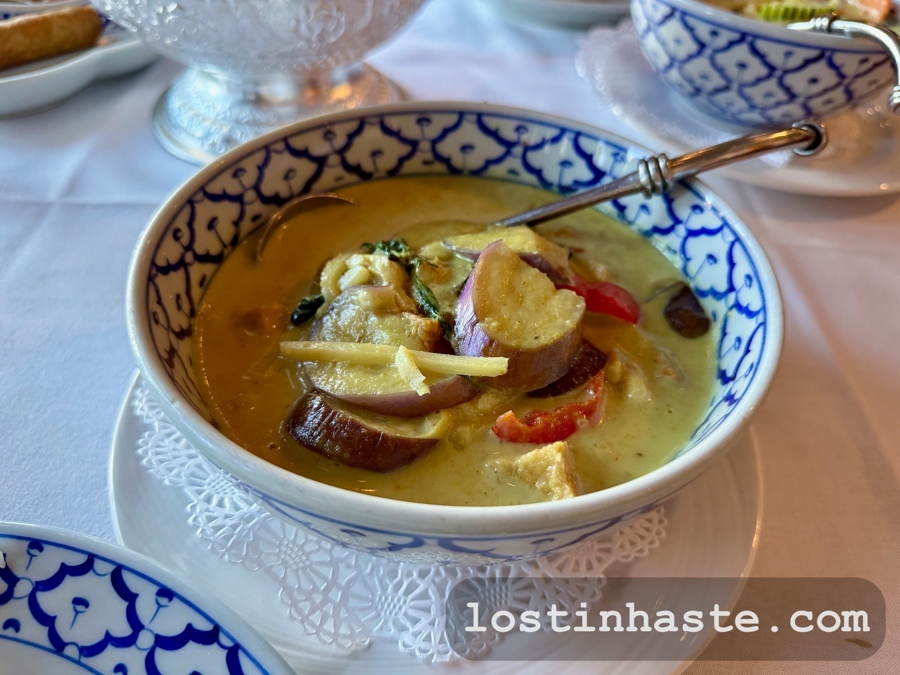 A bowl of green curry with vegetables and tofu is placed on a table, accompanied by a spoon.