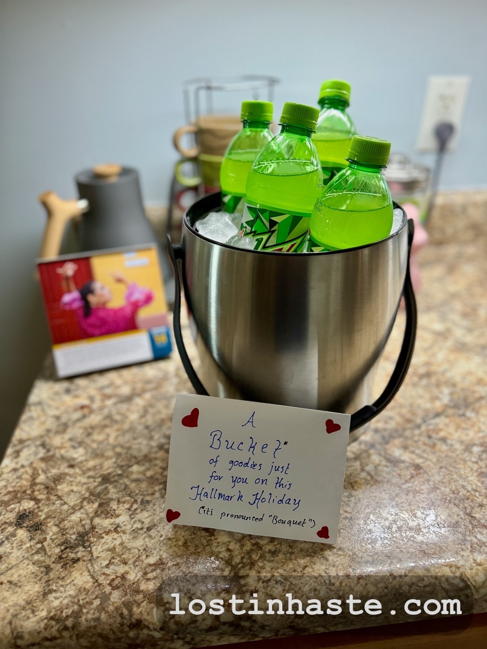 A metal bucket filled with ice and green bottled beverages on a countertop with a note, with heart stickers, reading: A Bucket* of goodies just for you on this Hallmark Holiday (*its pronounced 'Bouquet')