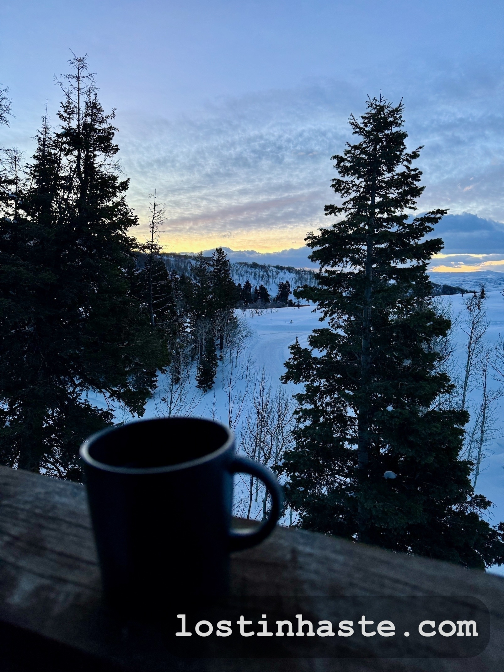 A mug rests on a wooden rail overlooking a snow-covered landscape with evergreen trees and a colorful sunrise sky. Text: lostinhaste.com