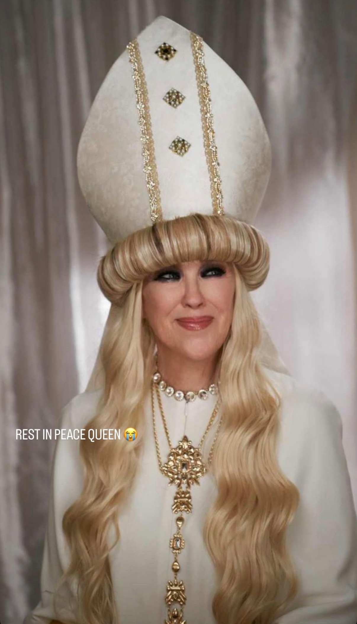 Auto-generated description: A woman dressed in ornate white robes with long blonde hair and a large headdress is smiling, accompanied by the text REST IN PEACE QUEEN 😢.