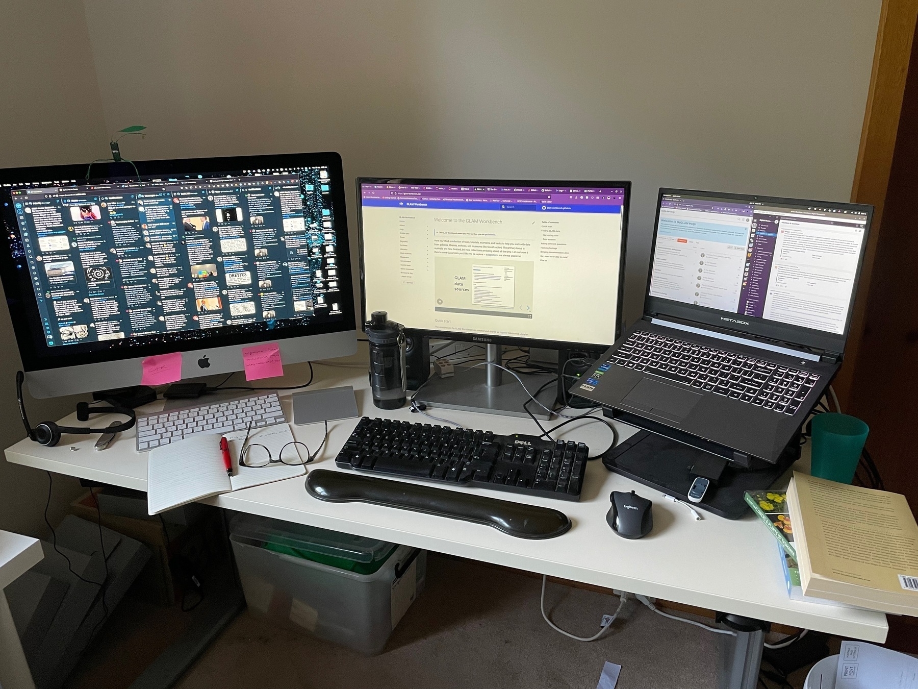 Photograph of my desktop. There are three monitors - one iMac, and a laptop on a stand connected to an external monitor. In the foreground are my distance glasses and a notebook.