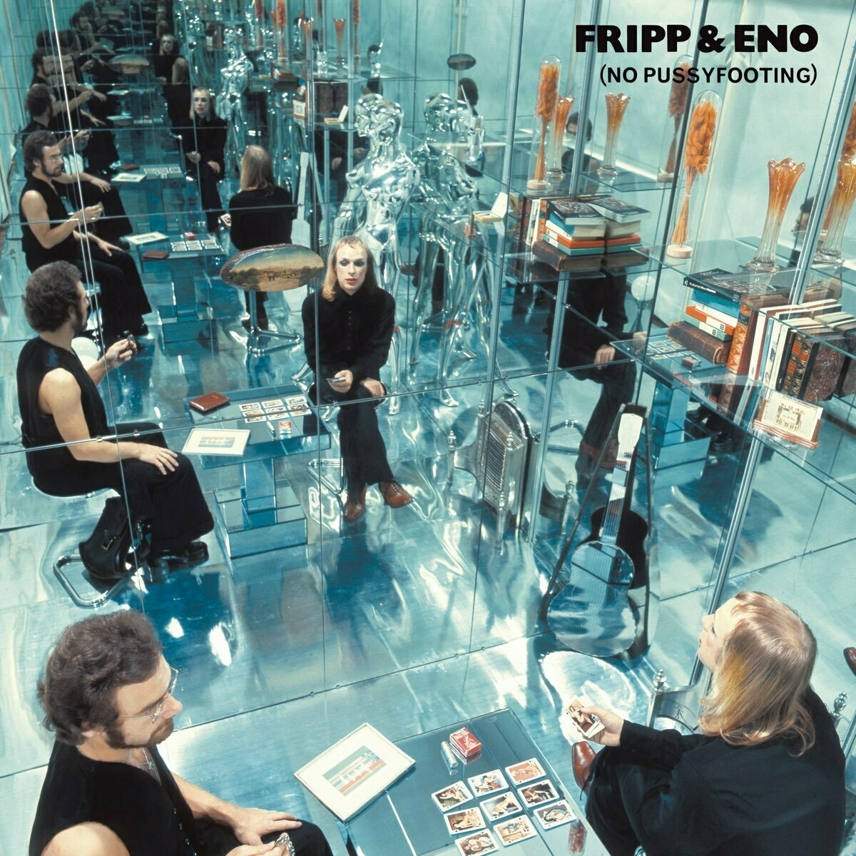 Album cover for Fripp & Eno: No Pussyfooting. Brian Eno is holding a deck of what may be tarot cards. Nine colorful cards are dealt on the table beside him, in three rows of three. There is a small framed painting lying nearby. Eno's pale hair is long and straight, and he is made up with eye shadow, lipstick, and blush. Robert Fripp, wearing glasses and with a neatly trimmed beard which matches his curly head hair, sits facing Eno. They are in a mirror-lined room, so their reflections repeat a dozen or more times into the distance, growing progressively dimmer. A glass shelf is visible over Eno's head, holding a small collection of old leather-bound books. There are many other elements in the small, shiny room that seem indescribable.