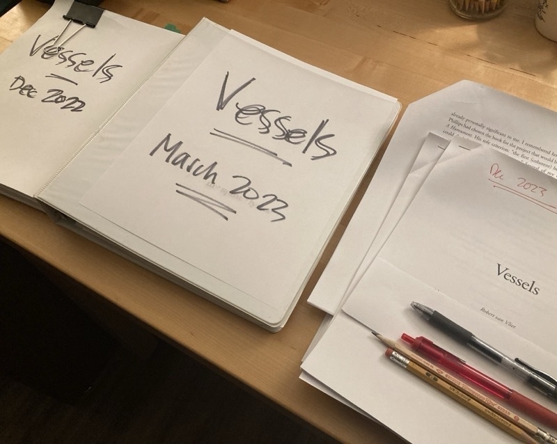 Three versions of a manuscript: One bound by a binder clip, titled Vessels December 2022; one in a 3-ring binder, titled Vessels March 2023, and a printout of the final galley proof as a series of small packets stapled together, titled Vessels with Dec 2023 written along the top.