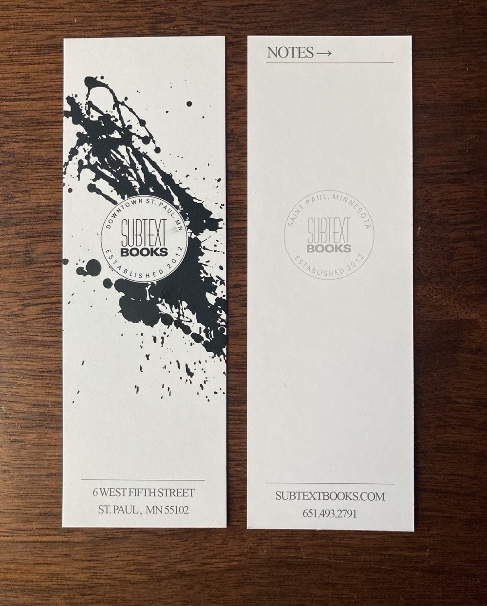 Two bookmarks from Subtext Books, one side showing a splattering of black ink, the other side largely blank but labelled Notes; in the center of both, a faint round stamp that reads: Subtext Books Downtown St Paul MN Established 2012