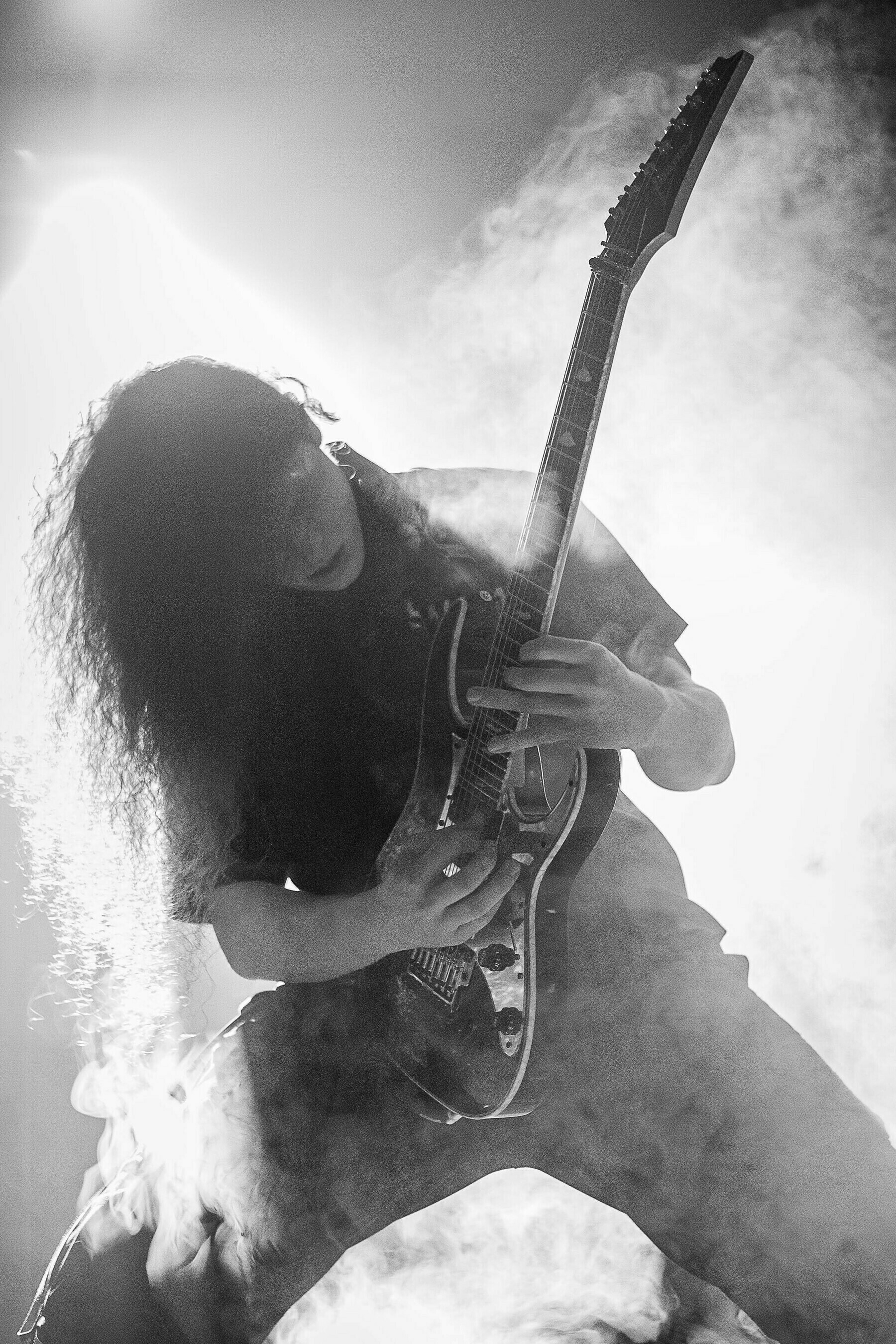 A man playing guitar in a heavy metal band with wild hair surrounded by lots of smoke.