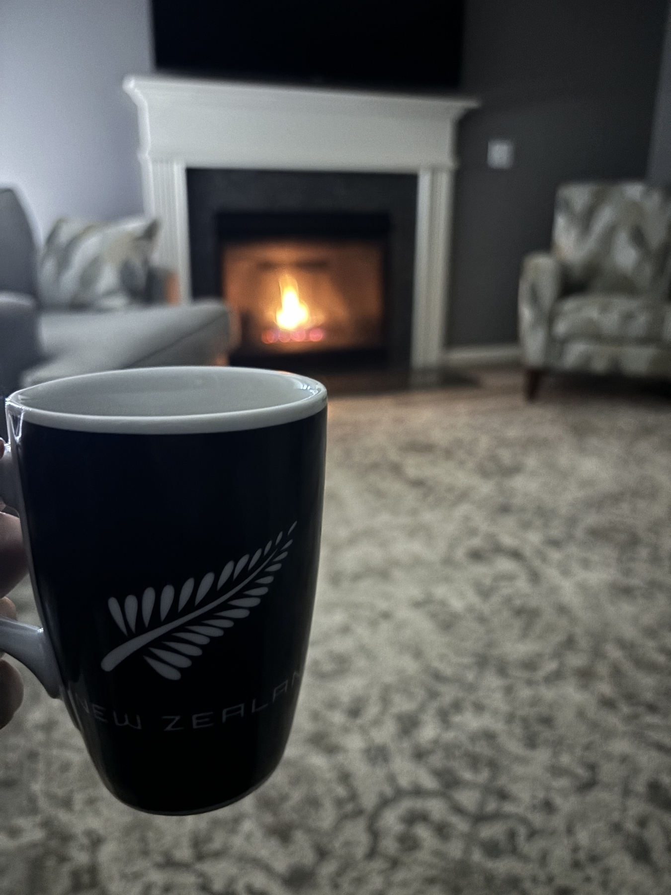 A coffee mug that reads, “New Zealand” in front of a fireplace with an inviting fire. 