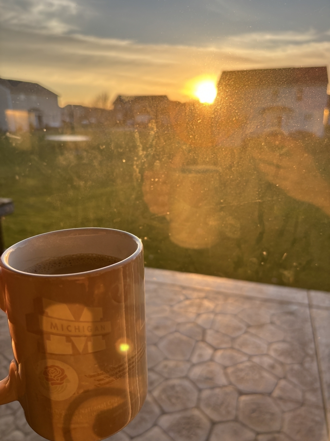 Coffee cup in the foreground while my reflection shows in a window looking over the sunrise. 