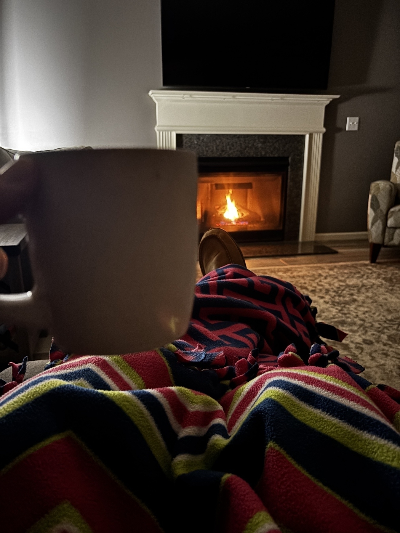 Cozy scene featuring a person relaxing with a cup of coffee, covered in a colorful blanket, and sitting by a lit fireplace.