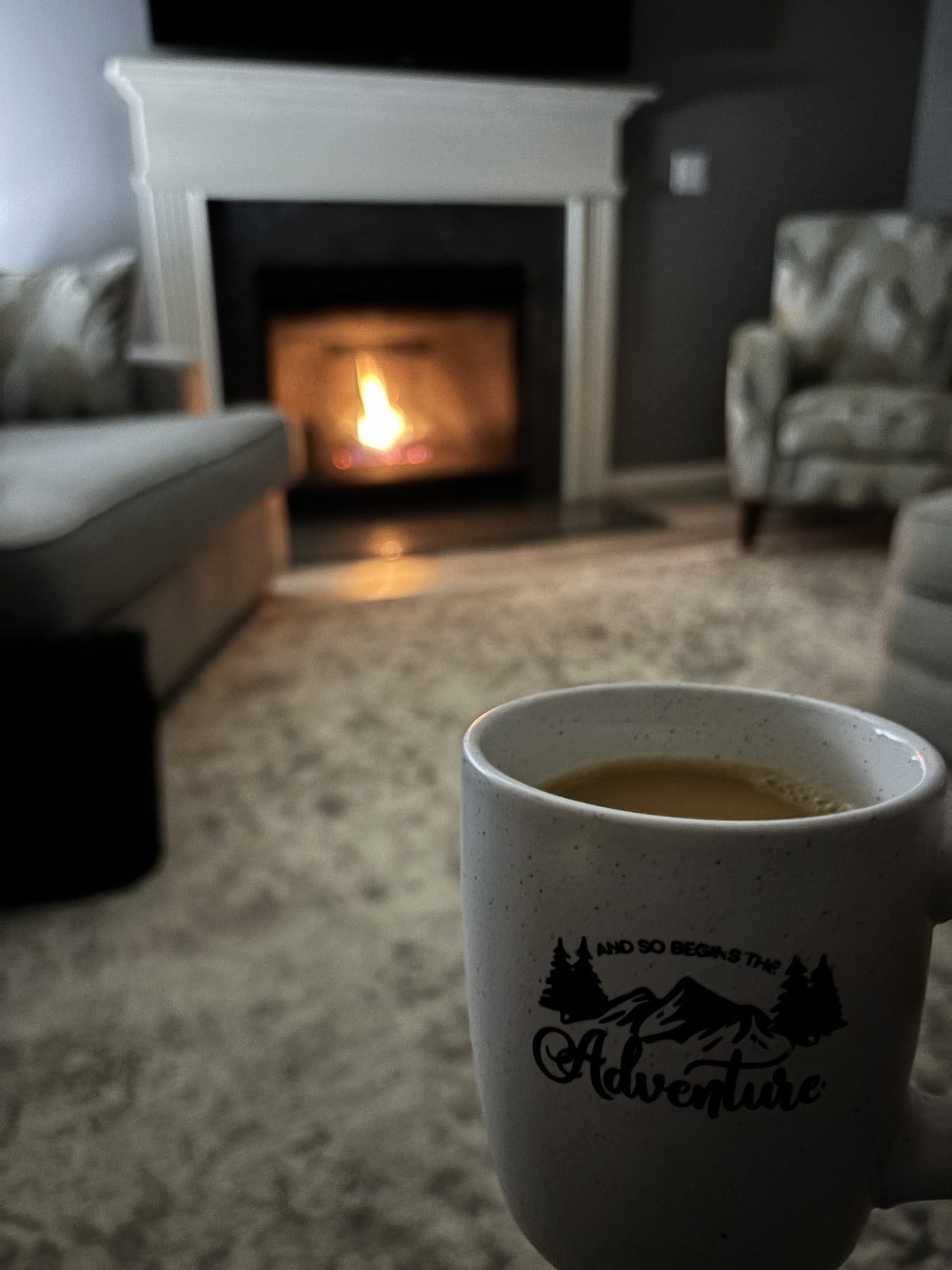 A coffee mug that reads, “Let the adventure begin” in front of a fire place