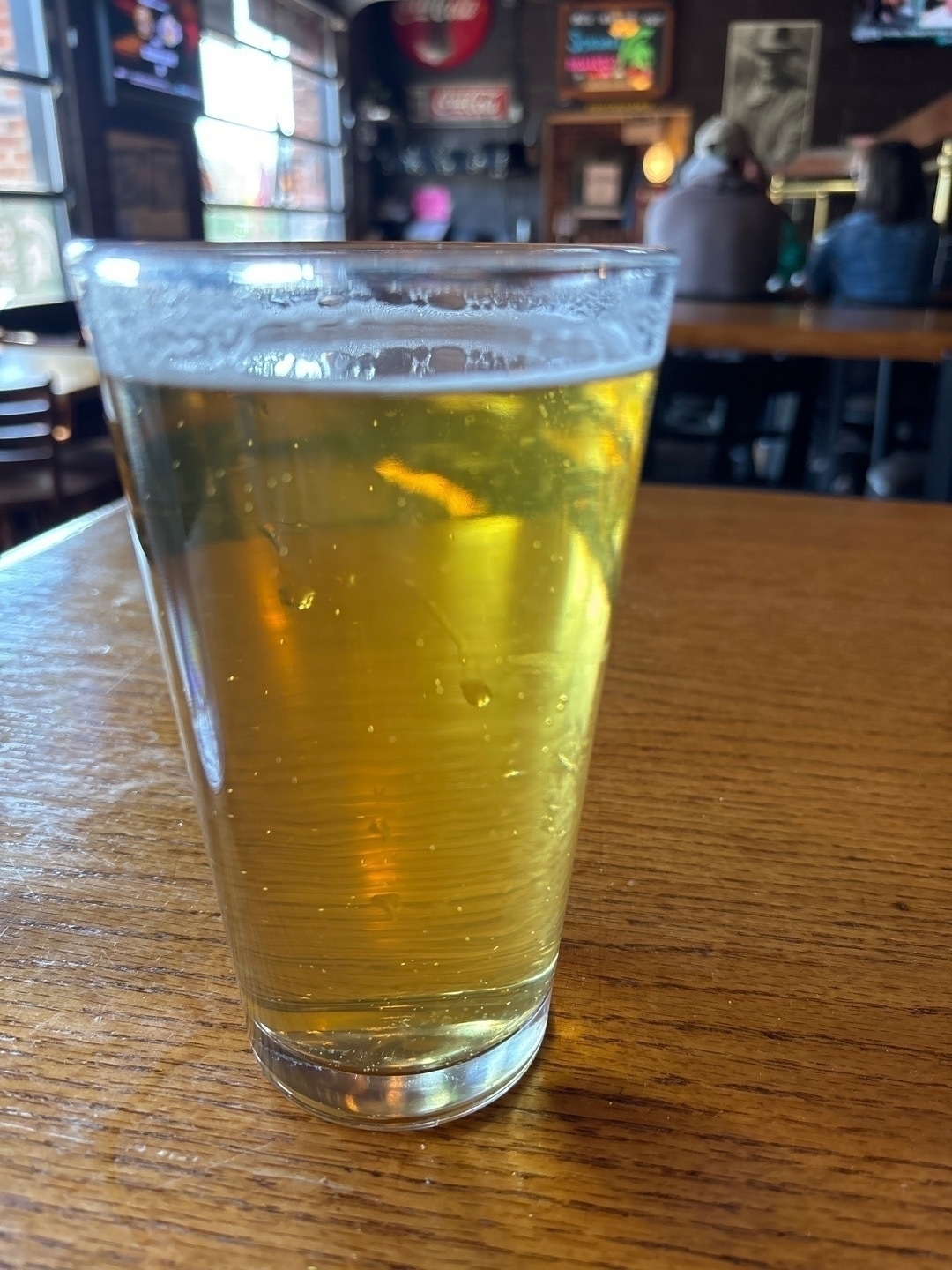 Auto-generated description: A glass of light beer sits on a wooden table in a bar setting.