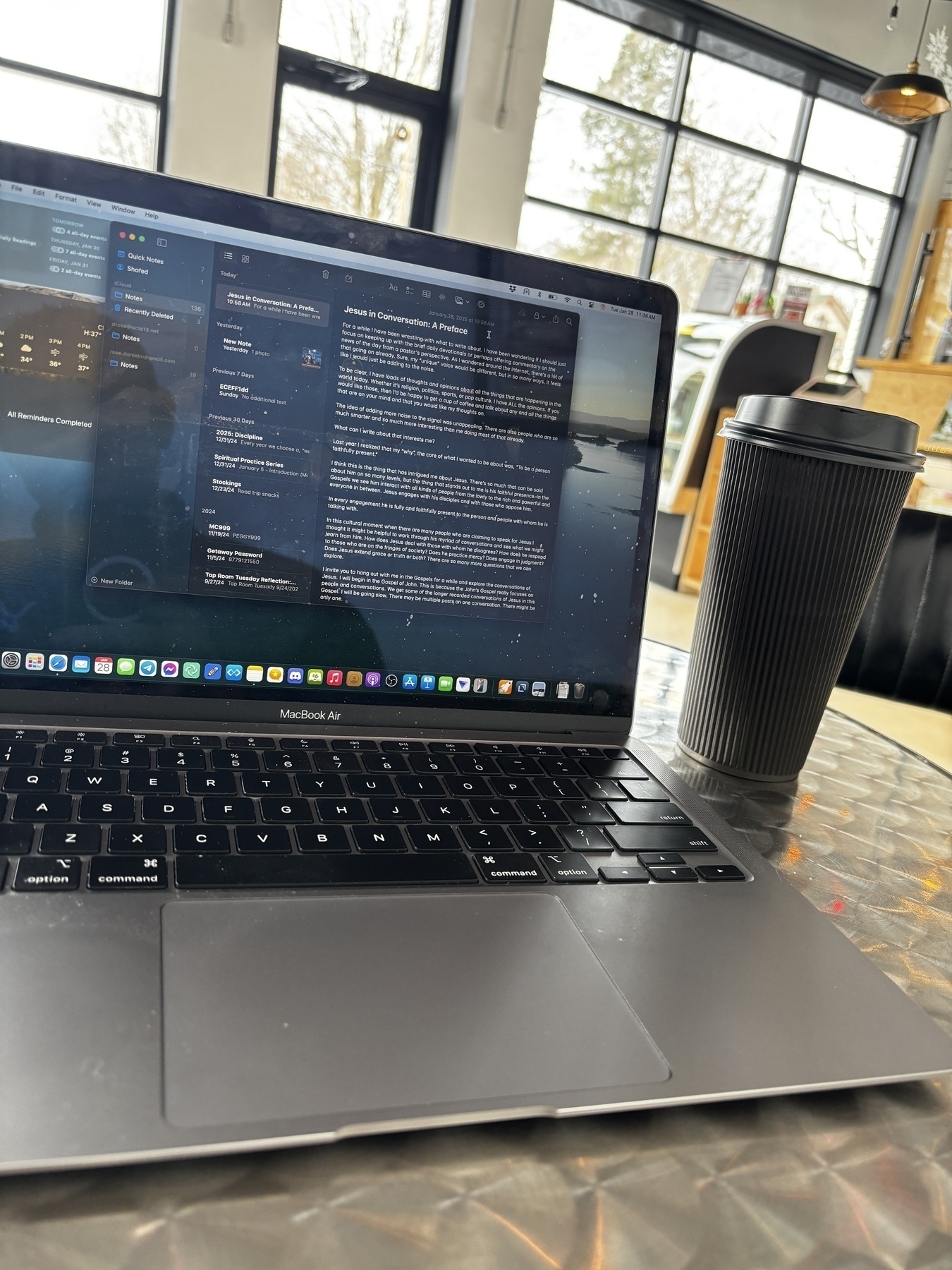 A laptop displaying a text document is placed on a metal table beside a black travel mug, with large windows and a door in the background.