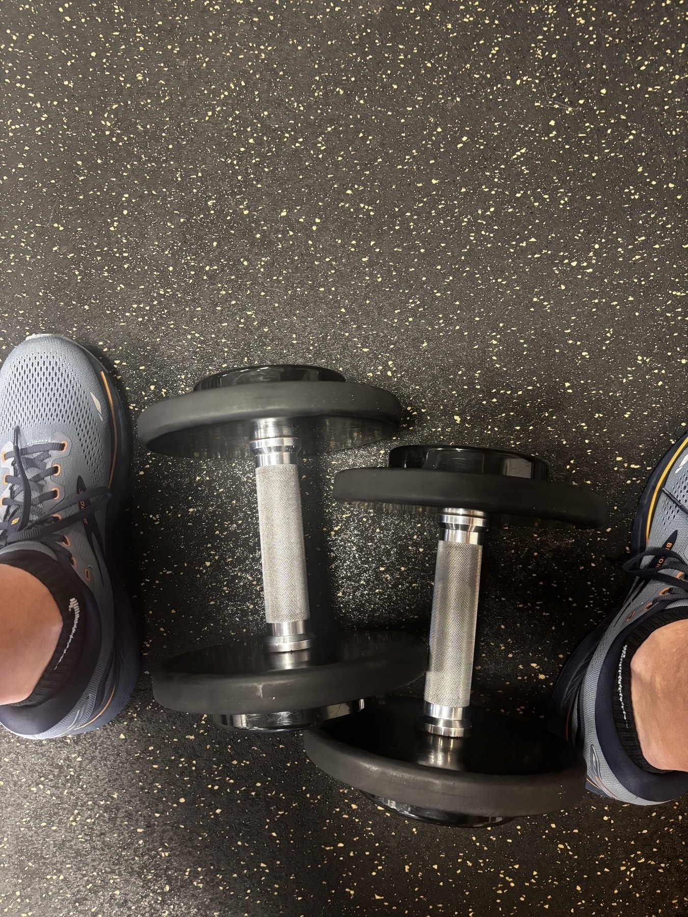 A pair of black dumbbells rests on a speckled gym floor near someone's feet wearing blue and gray athletic shoes.