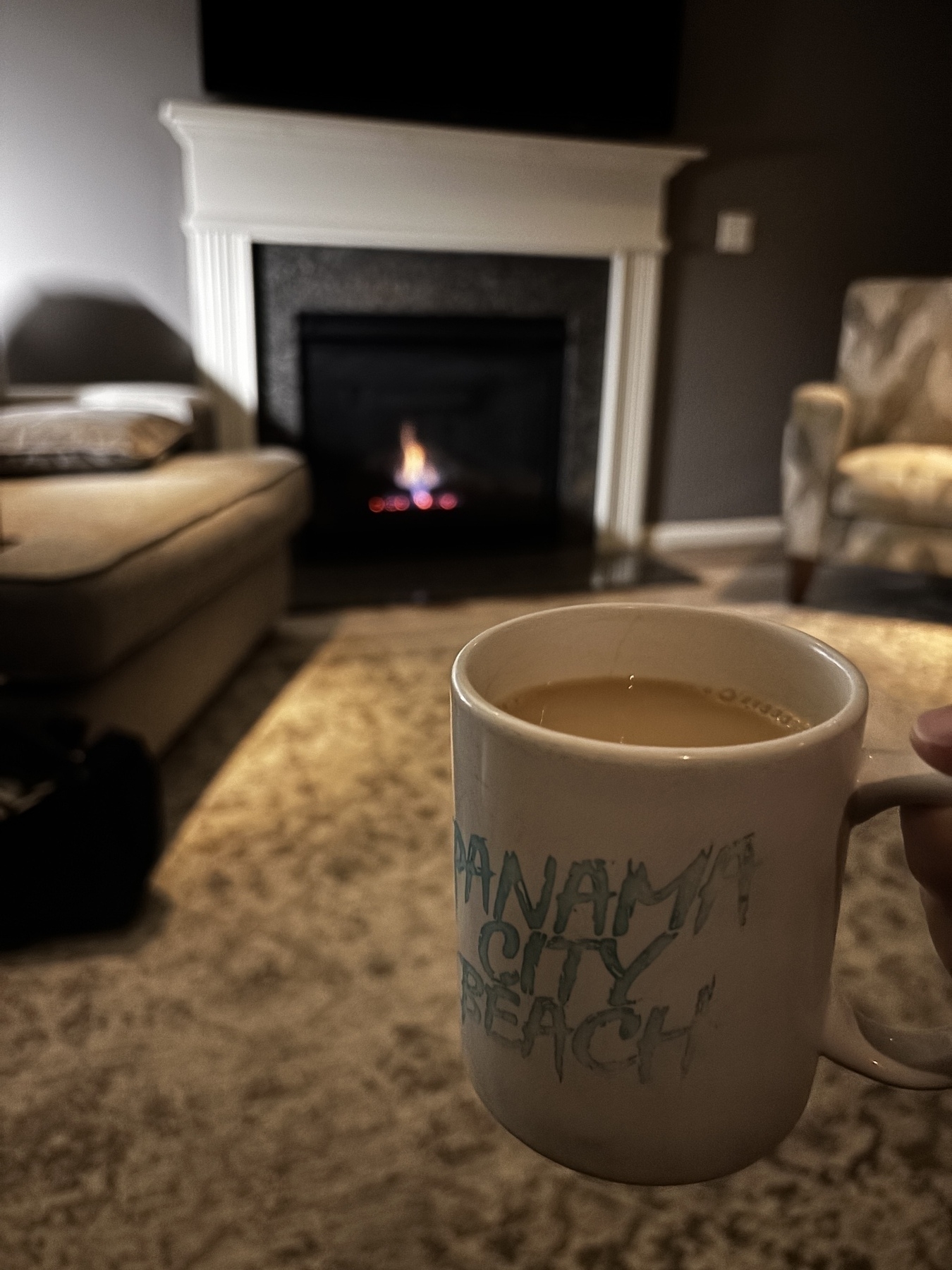 A cozy living room setting features a lit fireplace, a sofa, an armchair, and a person holding a coffee mug labeled "PANAMA CITY BEACH."