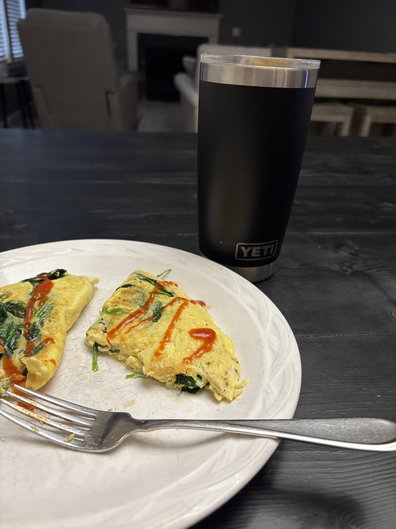 A plate with a partially eaten omelette topped with spinach and hot sauce is next to a black tumbler on a dark wooden table.