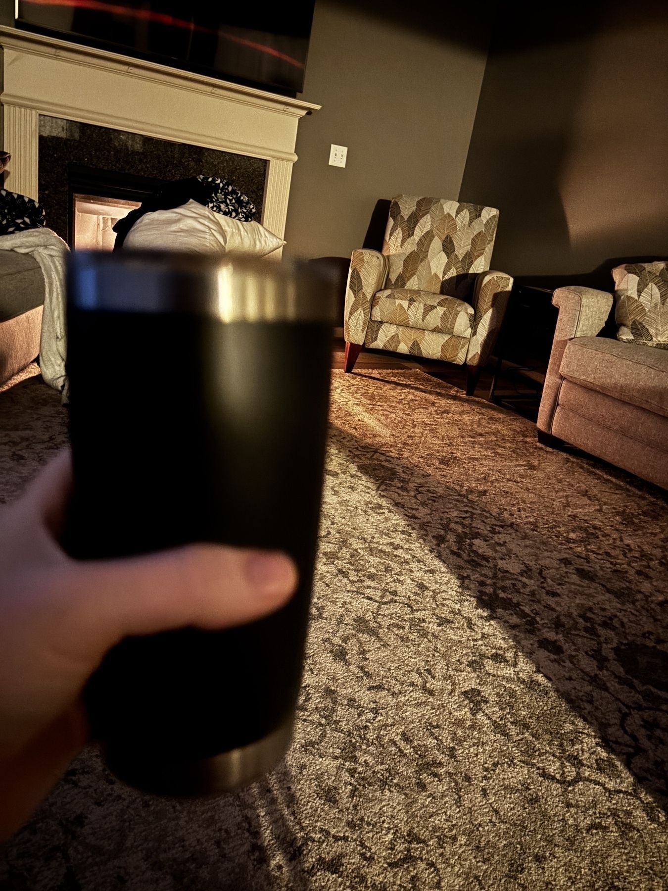 A person holding a black tumbler in a cozy living room with patterned armchairs and a fireplace.