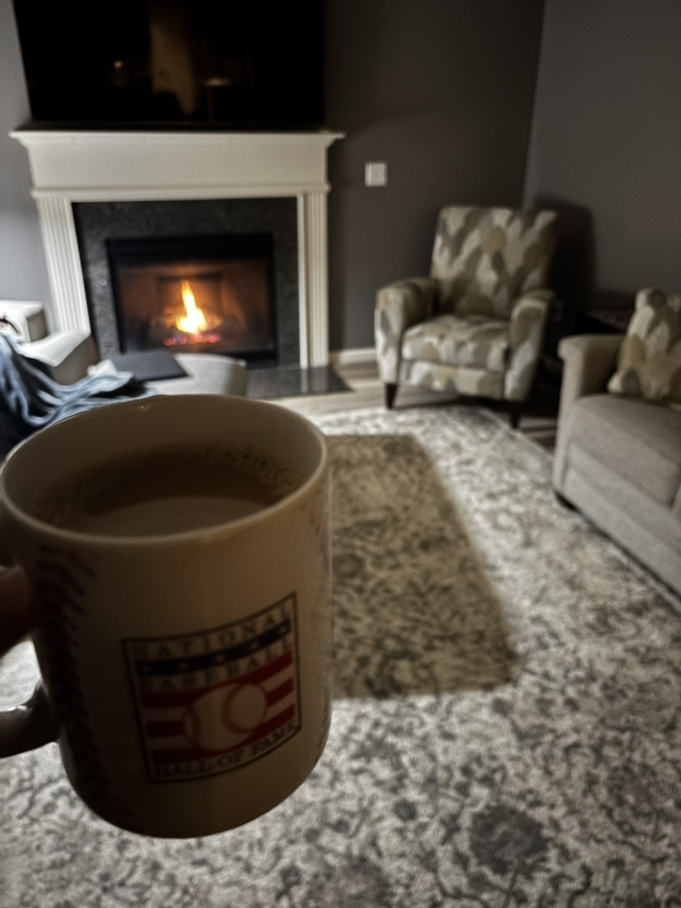 A person holds a coffee mug in a cozy living room with patterned chairs and a lit fireplace.