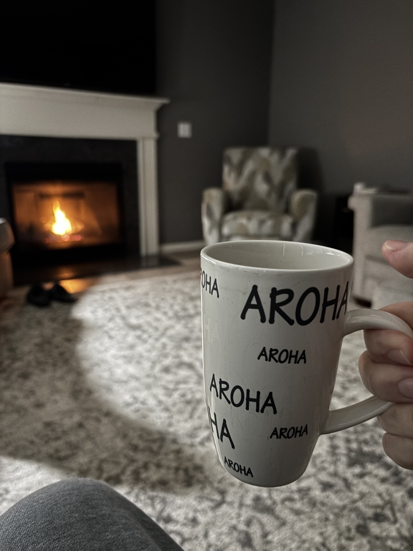 A hand is holding a mug with "AROHA" printed on it in a cozy living room with a fireplace and an armchair in the background.