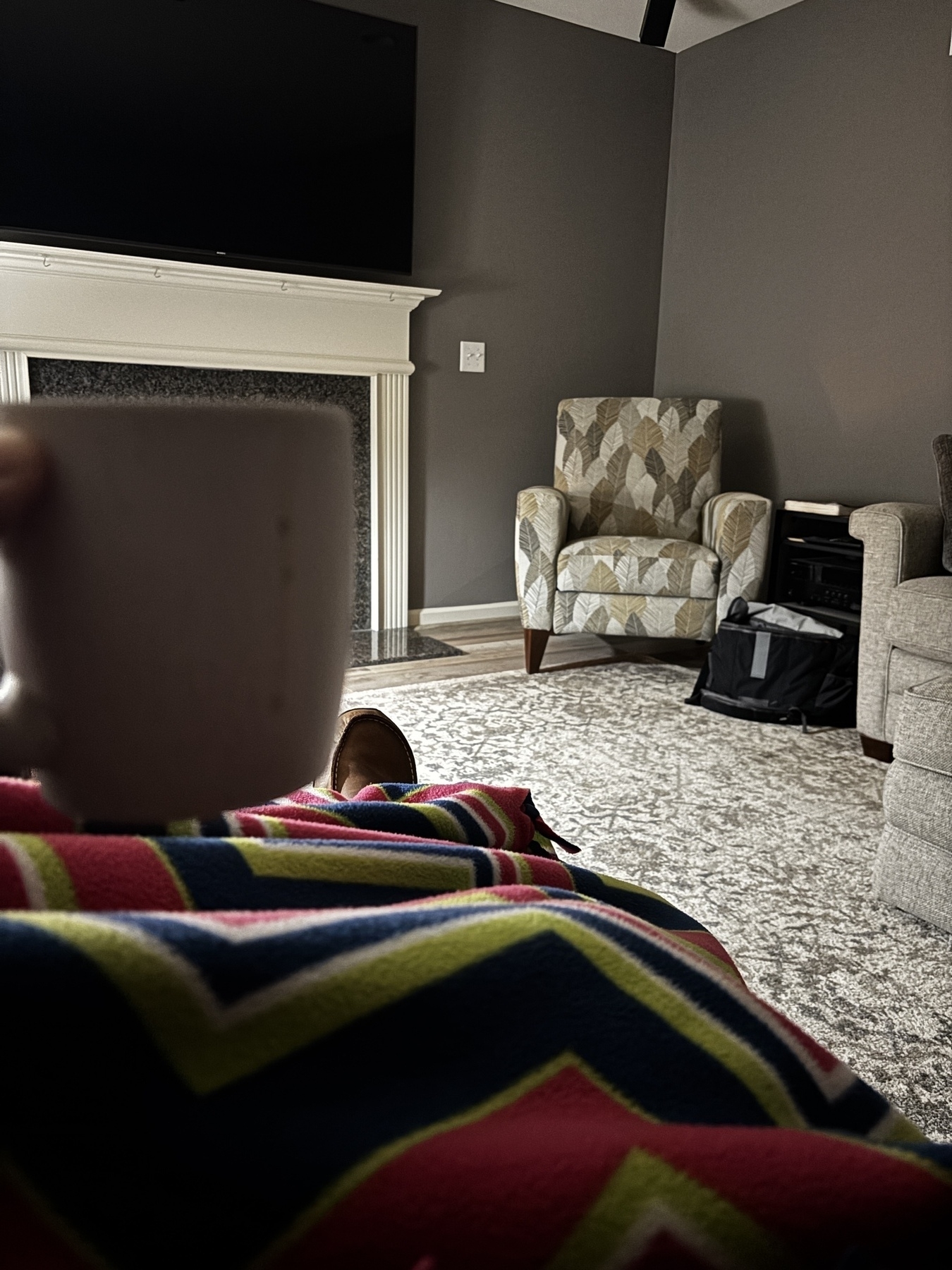 A person is sitting on a couch covered with a colorful blanket, holding a cup, in a living room with a fireplace and patterned armchair.