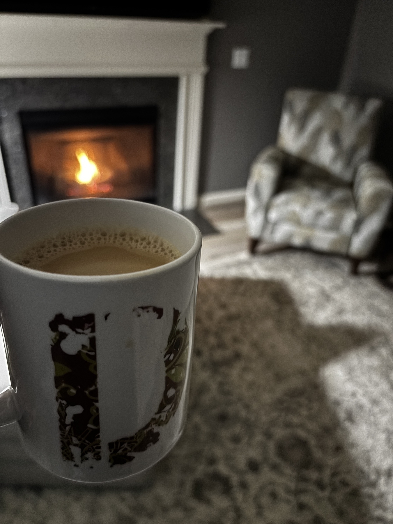 A mug of coffee is held in front of a fireplace with a patterned armchair nearby.