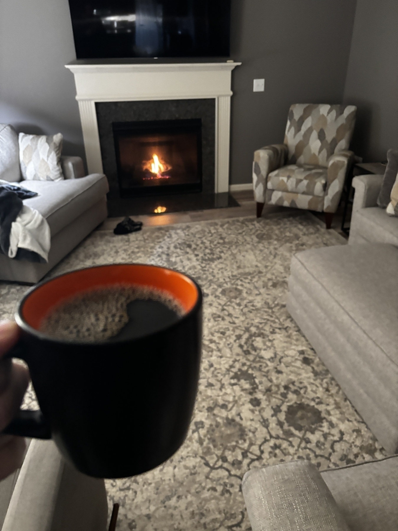 A hand holds a coffee mug in a cozy living room with a fireplace, chair, and sofas.