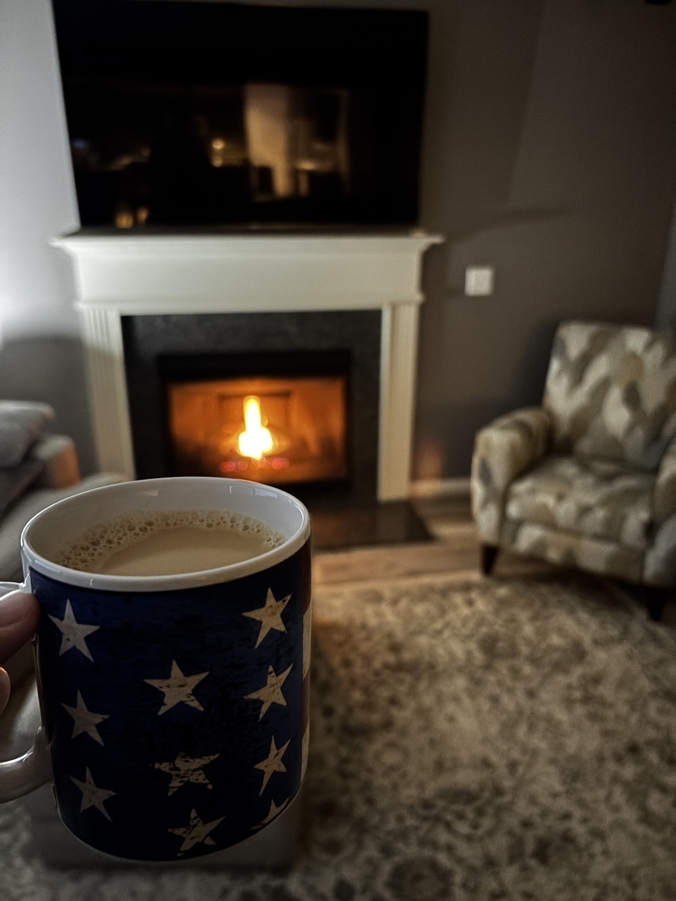 A cozy living room scene features a hand holding a star-patterned mug near a lit fireplace with an armchair to the right.