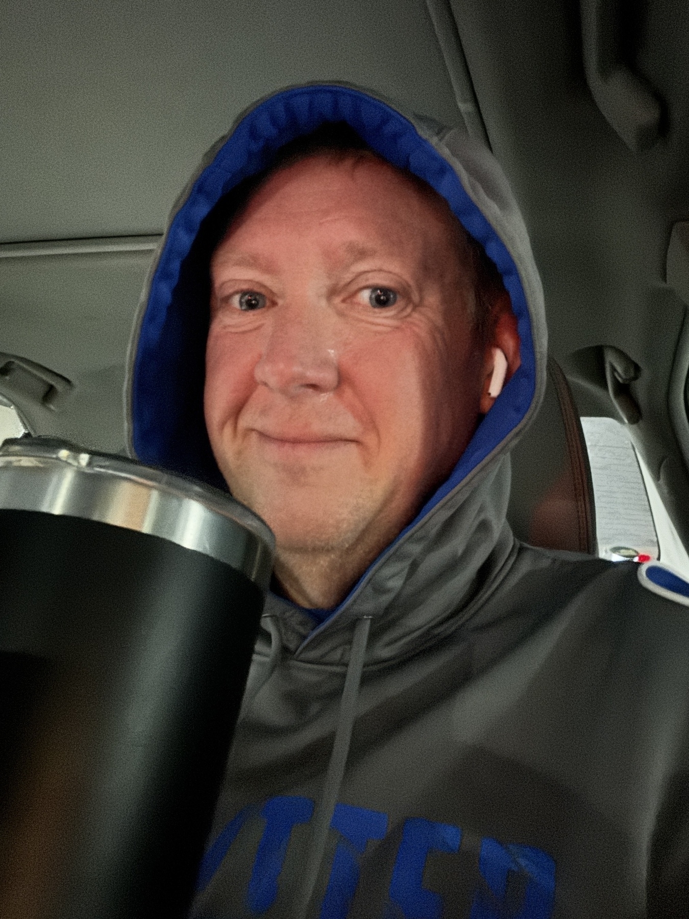 A person wearing a gray hoodie and AirPods holds a large black tumbler while sitting inside a vehicle.